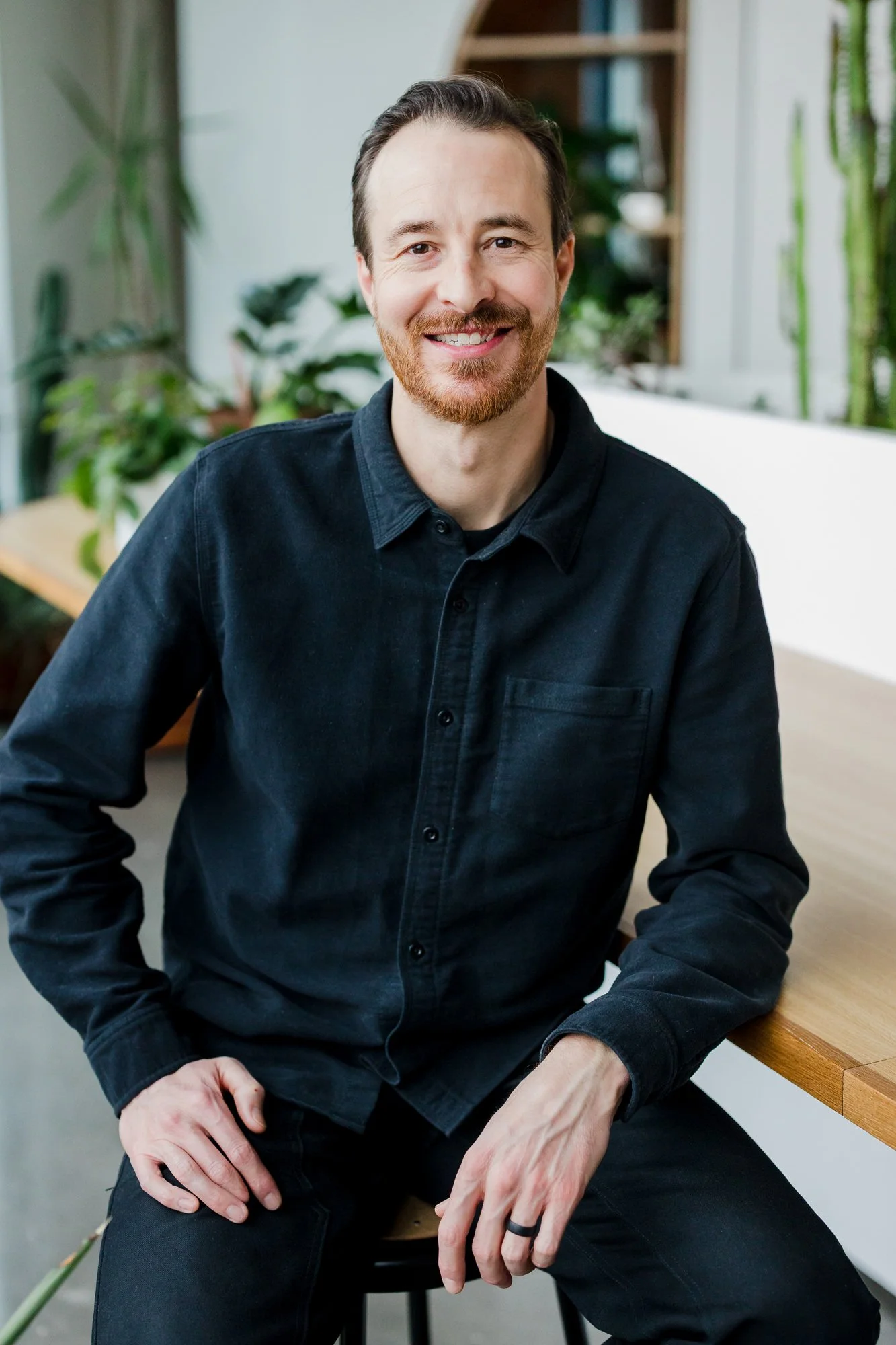 A man with a beard and mustache, wearing a black button-up shirt, sitting on a stool in a room filled with green plants, smiling at the camera.