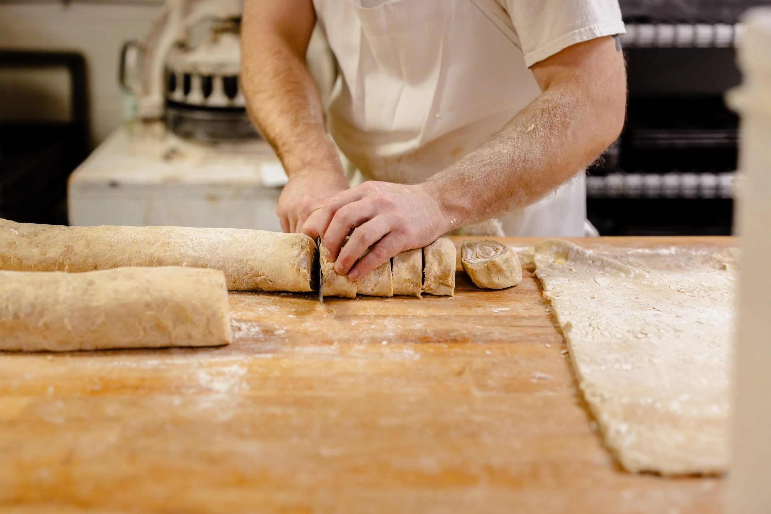 A person wearing a white apron slicing rolled dough into smaller pieces on a floured wooden surface, with more dough and baking tools around.