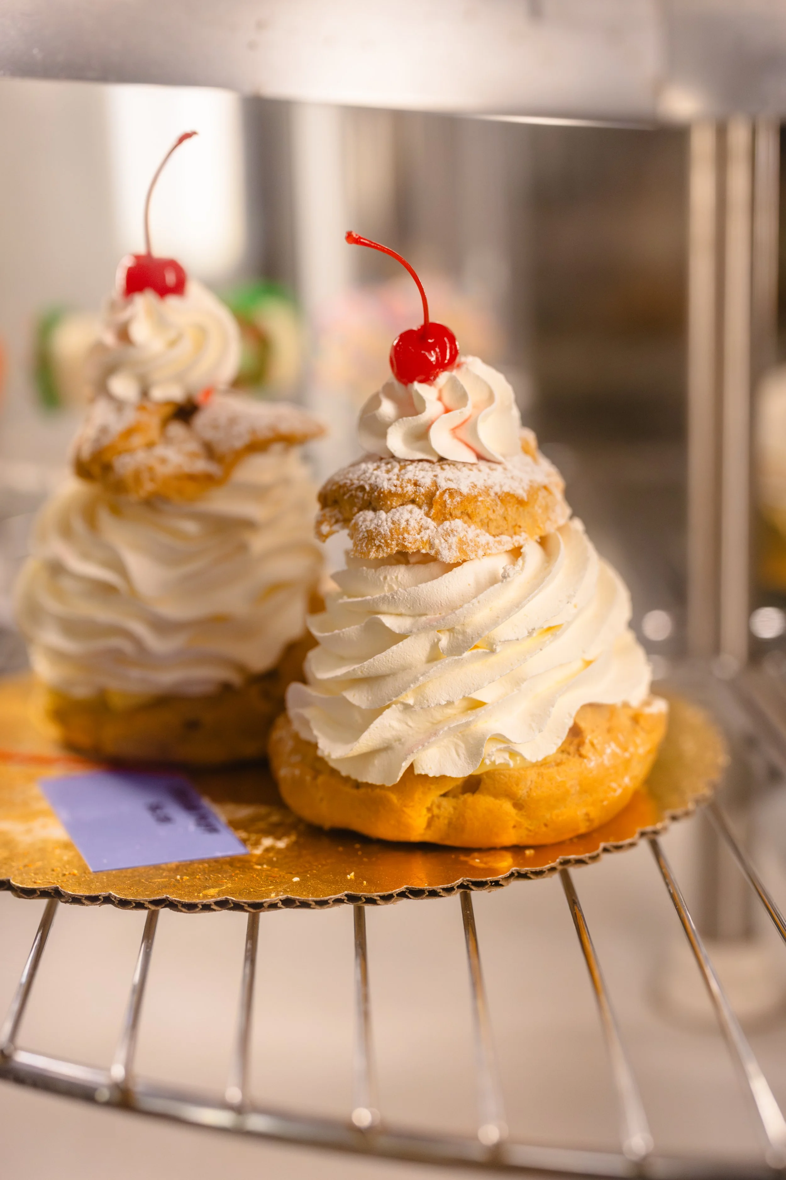Two cream puffs topped with whipped cream, a cherry, and a small cookie on a golden paper base, displayed on a cooling rack.