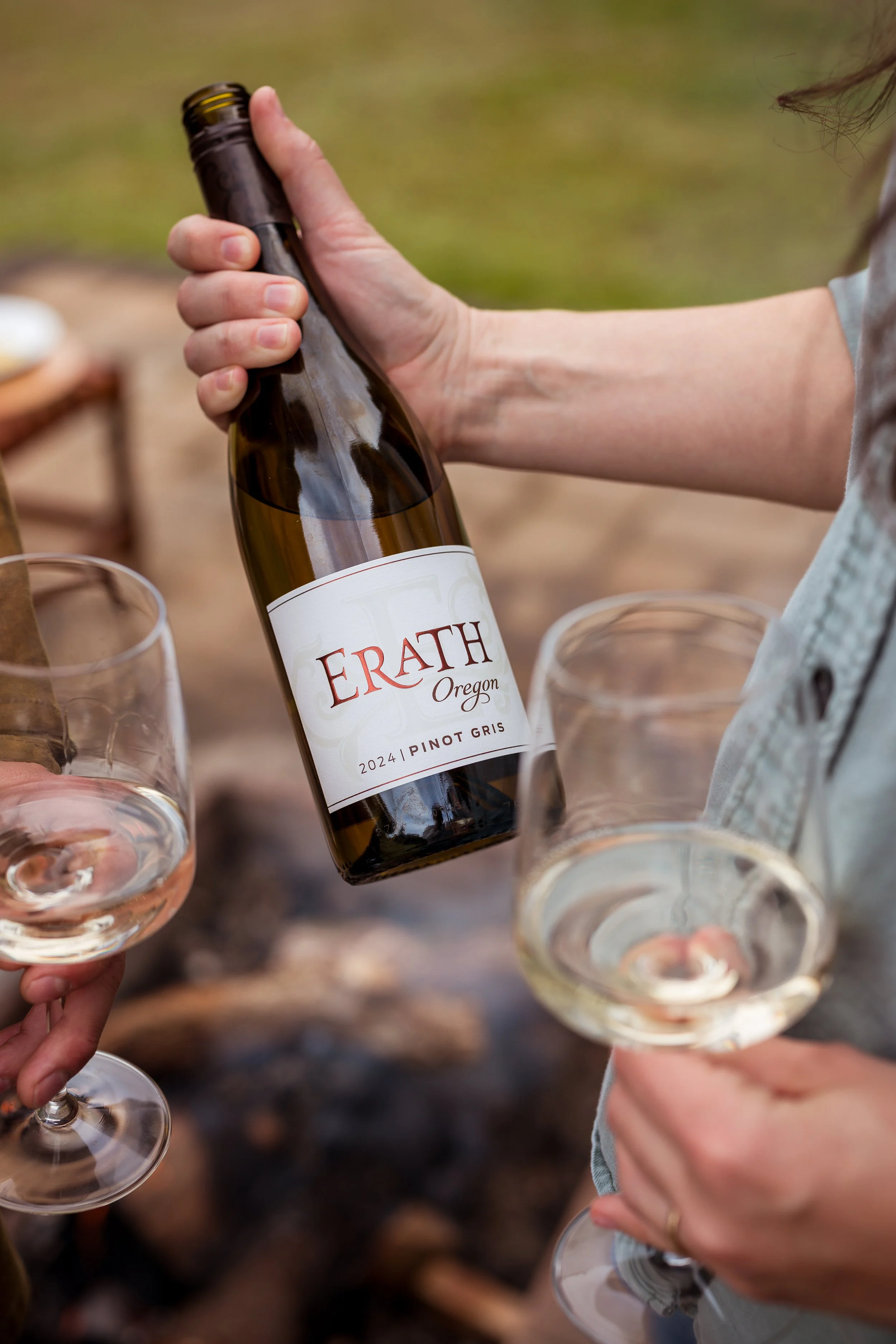 Person holding a bottle of Erath Oregon Pinot Gris wine, with glasses of white wine around, outdoors on a natural setting.