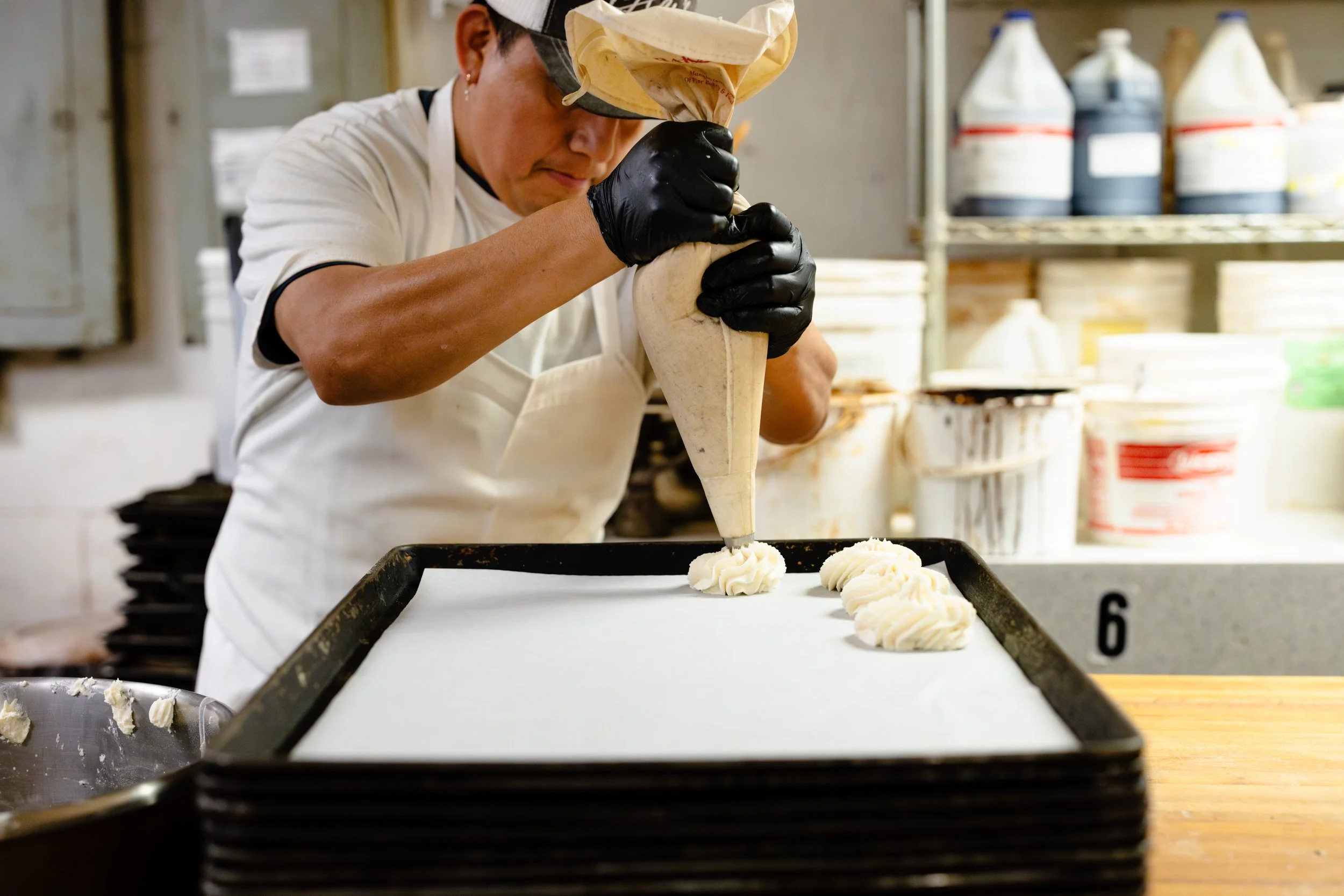 A baker wearing black gloves, a white apron, and a white cap is piping frosting onto cookies on a baking sheet.
