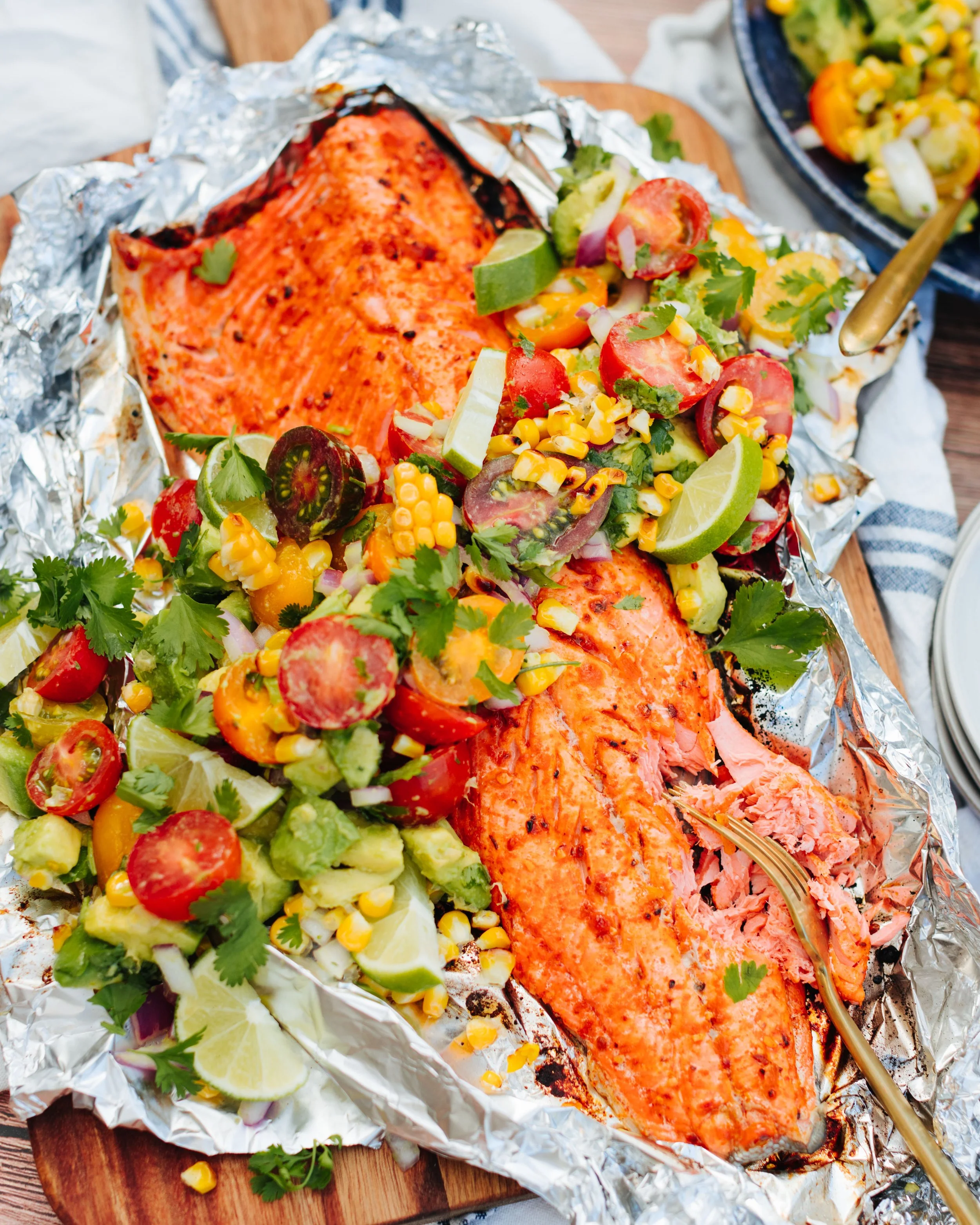 Baked salmon fillets topped with fresh tomato, corn, cilantro, and lime wedges, served on crinkled aluminum foil on a wooden platter, with a side of vegetable salad in the background.