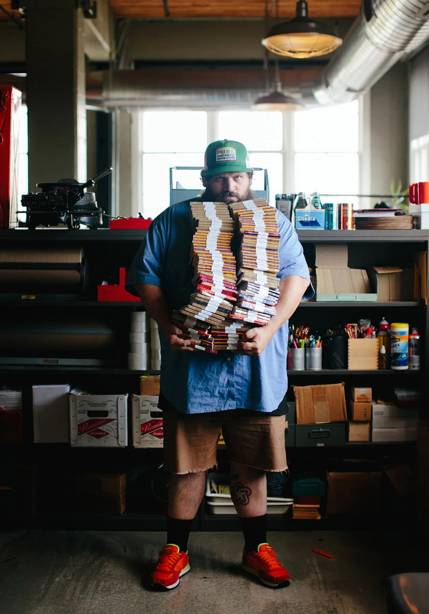 A man wearing a green cap, blue shirt, and red sneakers standing indoors holding a large stack of comic books or magazines, with shelves of art supplies and boxes in the background.