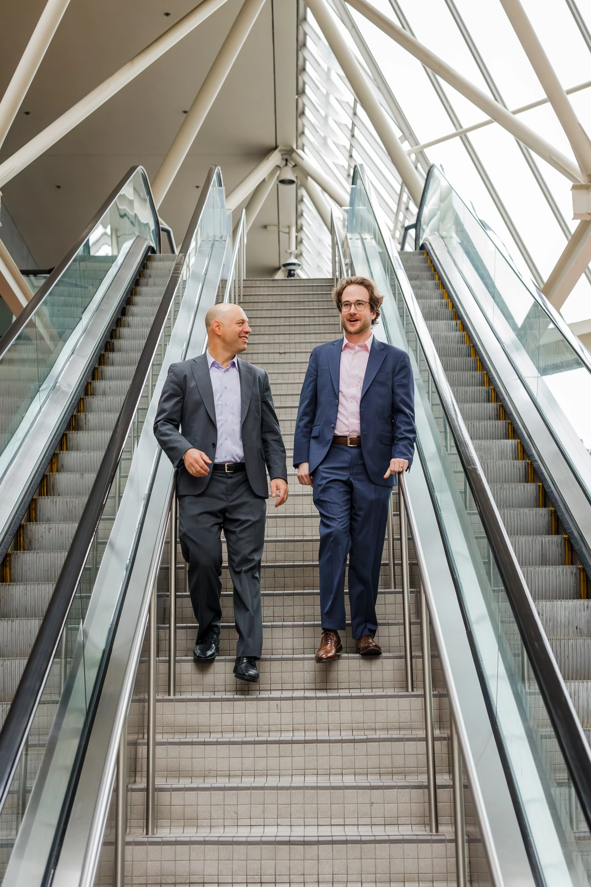 Two men in business suits walking down escalator stairs in a modern building with large glass windows.
