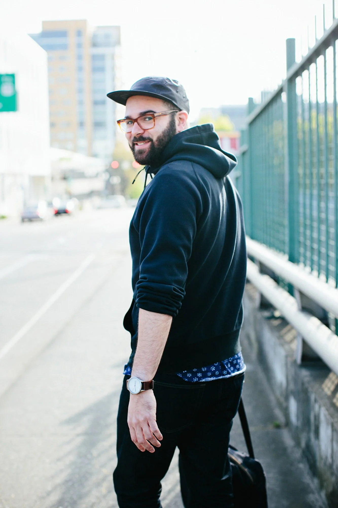 A man with a beard wearing glasses, a black cap, a black hoodie, and dark jeans, standing on the sidewalk near a green fence with a cityscape in the background.
