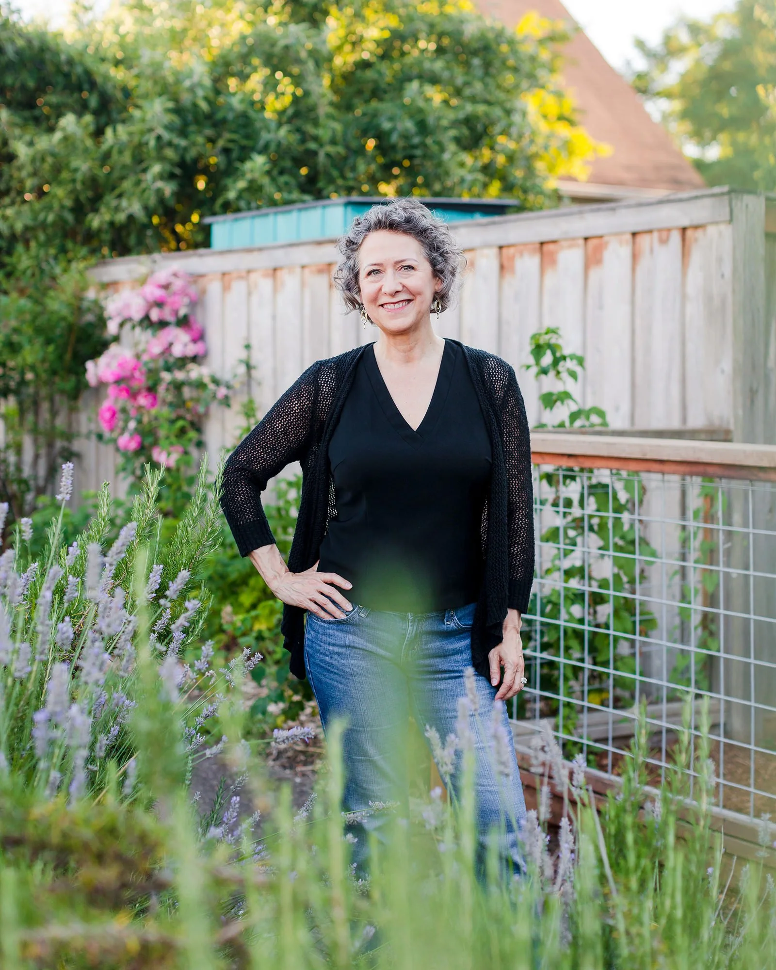 A smiling woman with curly gray hair standing outdoors in a garden with blooming flowers and greenery, wearing a black top, a black cardigan, and blue jeans.