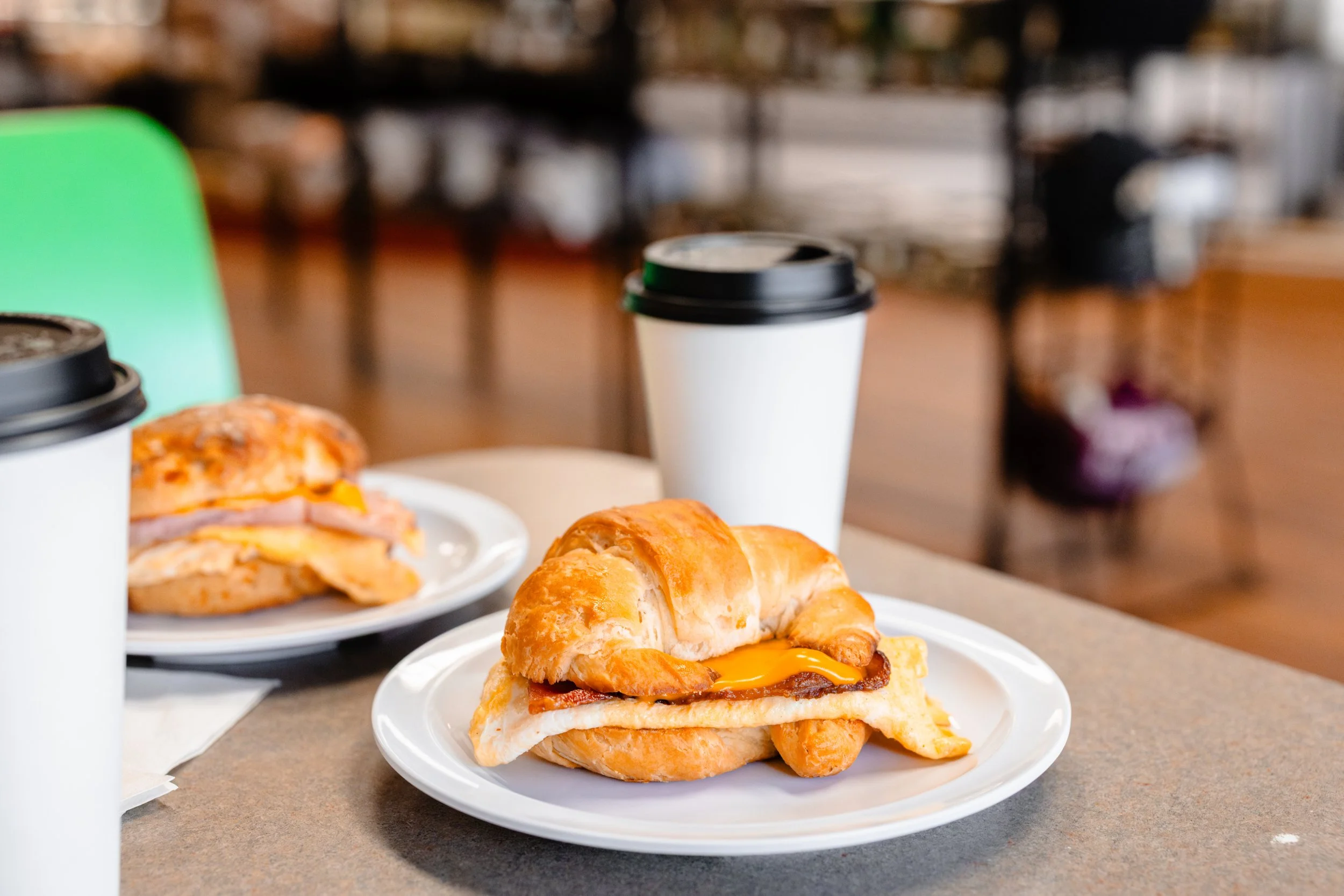 A croissant sandwich with egg and bacon on a white plate, two cups of coffee with black lids, and a second sandwich on a plate in the background. The setting appears to be a breakfast cafe with a blurred background.