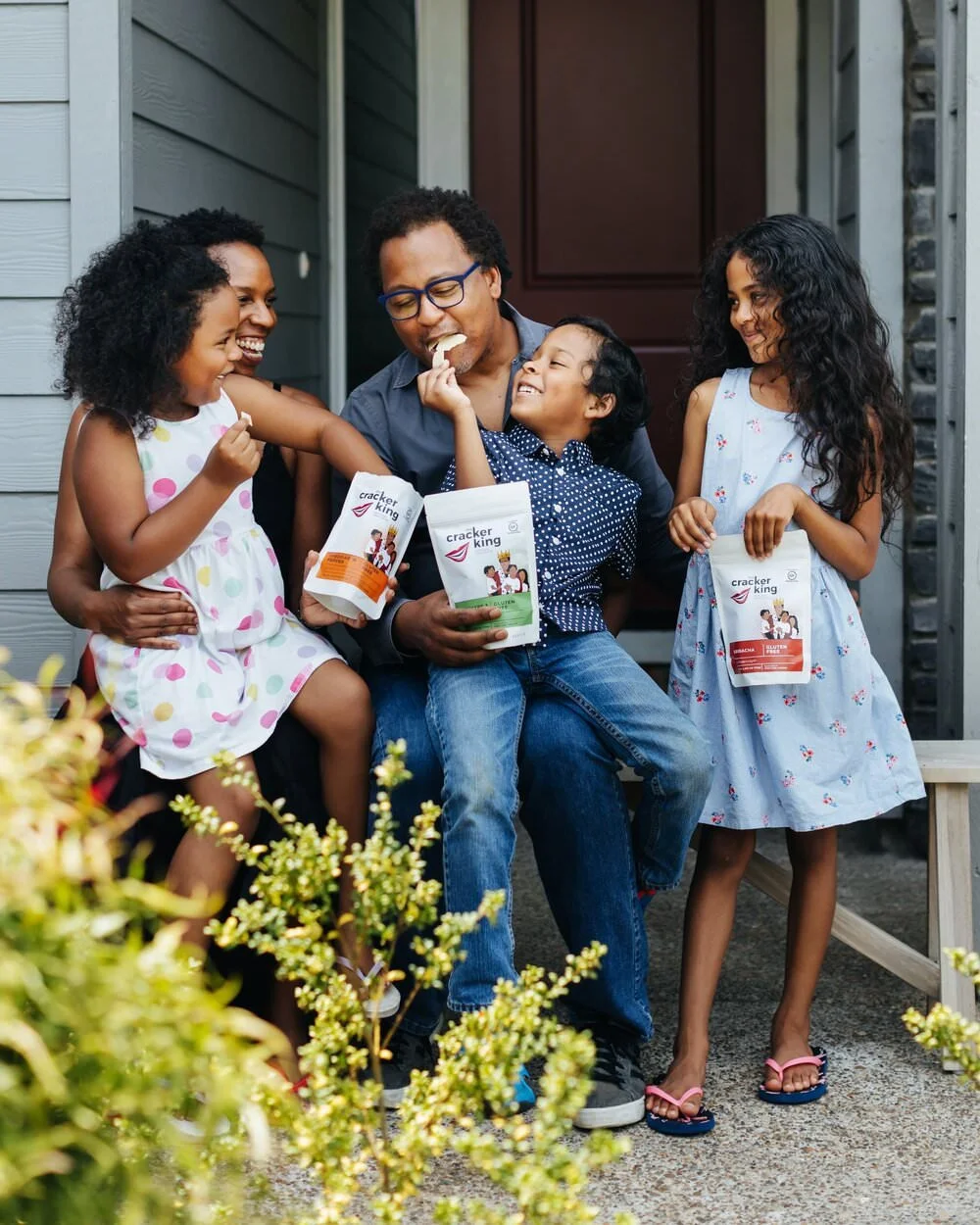 A family of five enjoying a moment together on the front porch, sharing crackers from the brand 'Cracker King', with two girls and one boy, all smiling and engaged.
