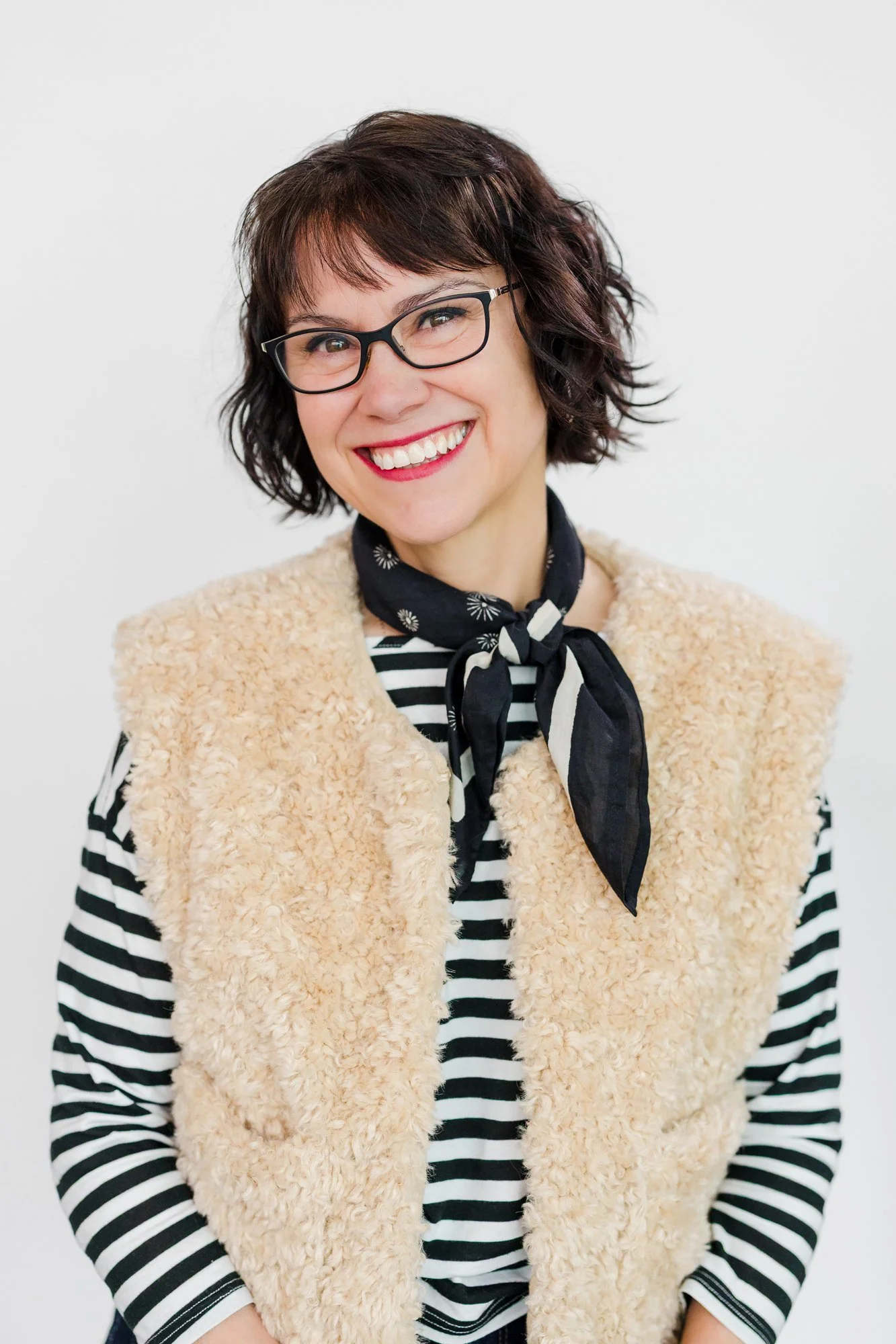 A woman with short, dark, curly hair, wearing glasses, a striped shirt, a black and white scarf, and a beige, fuzzy vest, smiling at the camera.