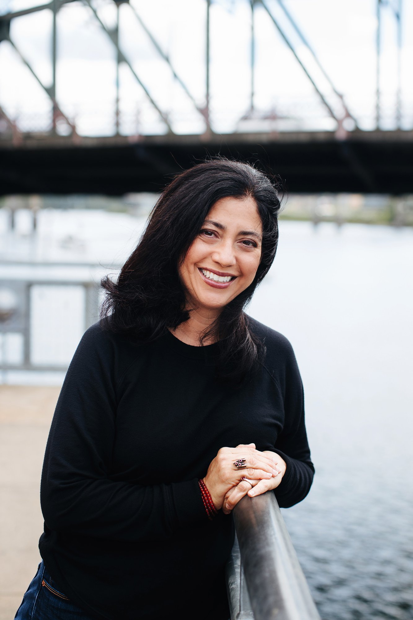 A woman with black hair and a black long-sleeved shirt smiling, standing outdoors near a river or body of water, holding a metal railing with her hands, with a bridge and boats in the background.