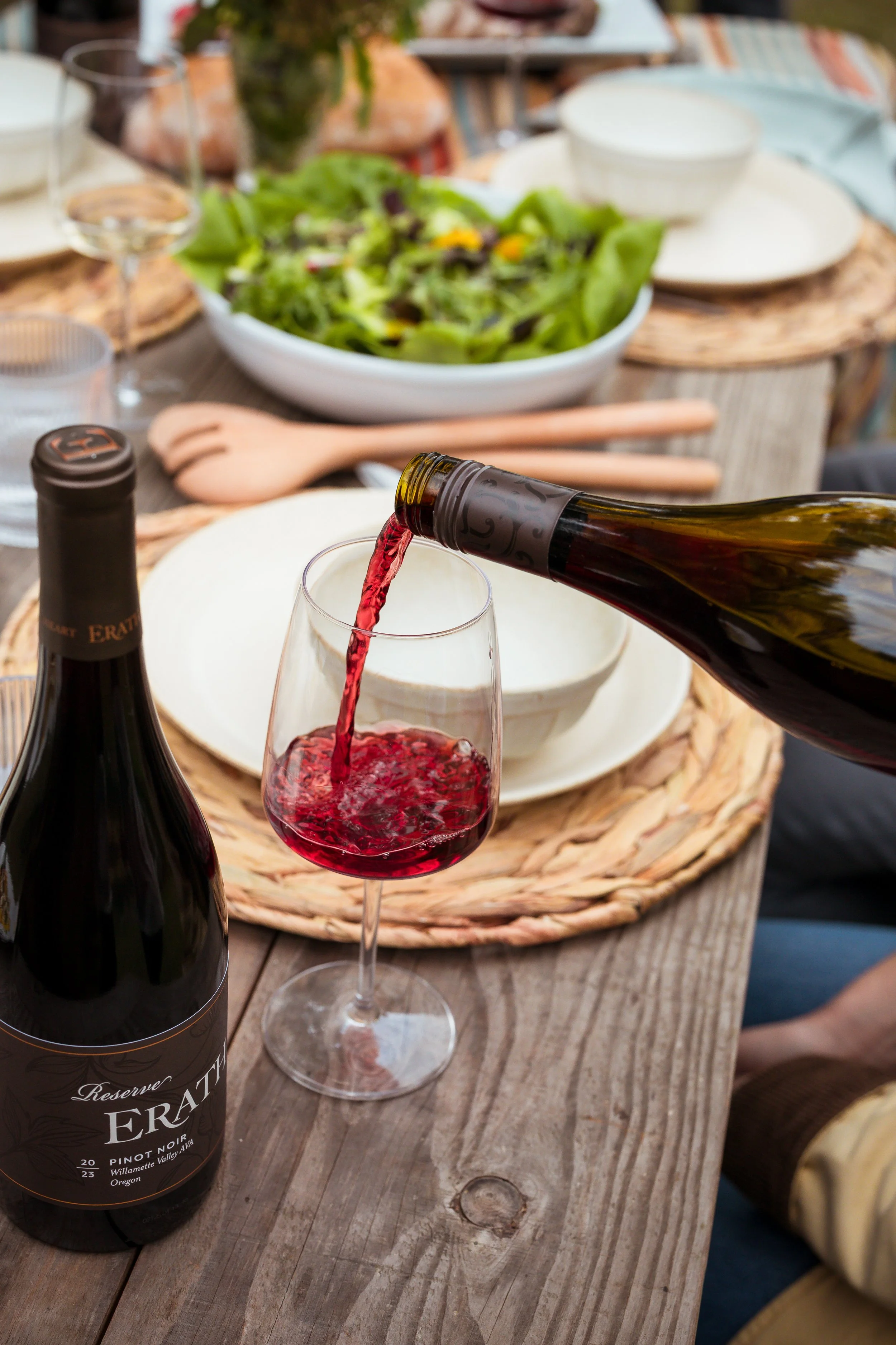 Pouring red wine into a wine glass at a rustic wooden table set with a salad, wine bottles, and plates for a meal.