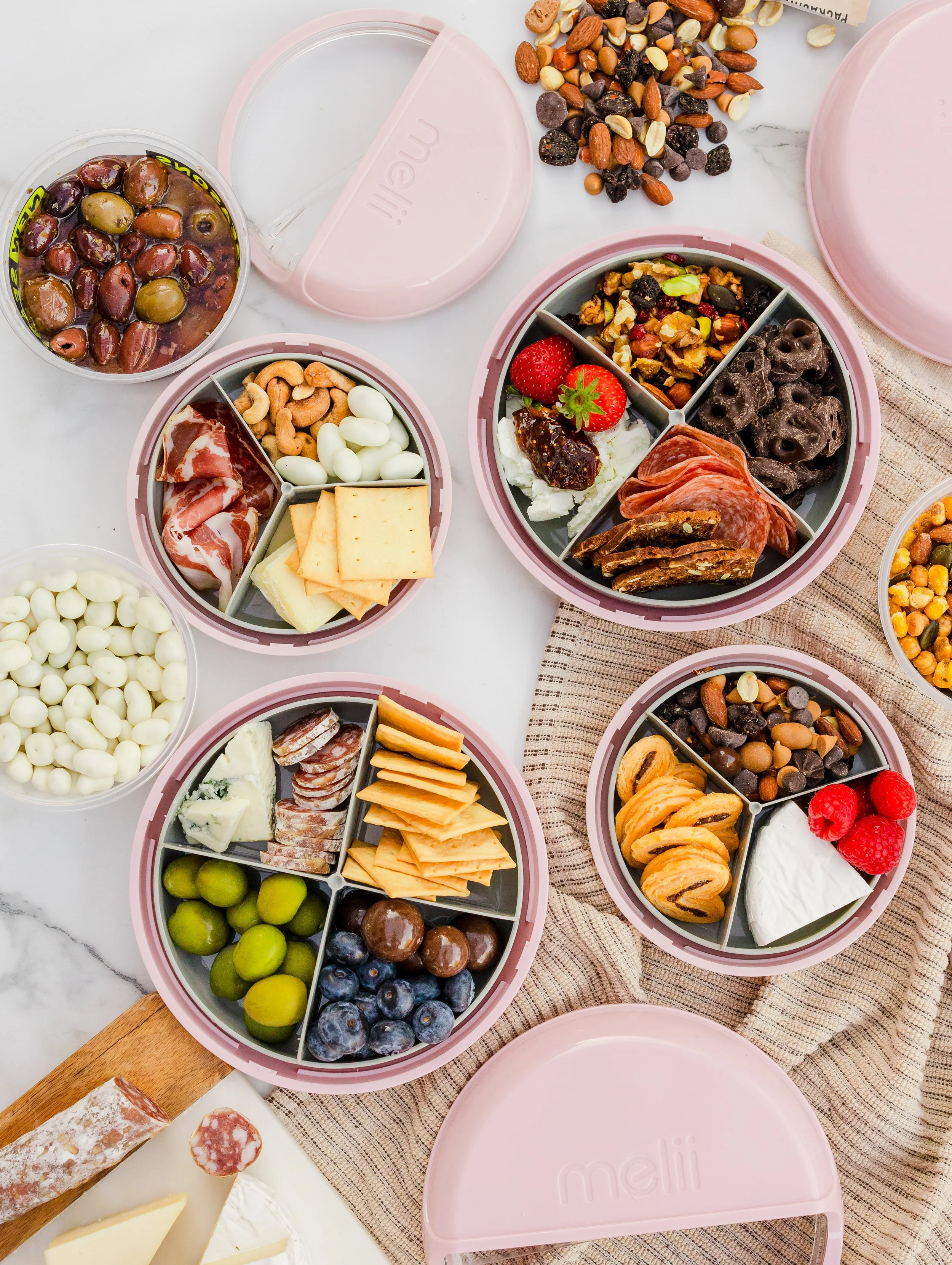 A variety of snacks and appetizers arranged in pink meli divided containers, including grapes, blueberries, olives, assorted cheeses, nuts, dried fruits, meats, chocolate-covered pretzels, strawberries, raspberries, and crackers, on a white marble su