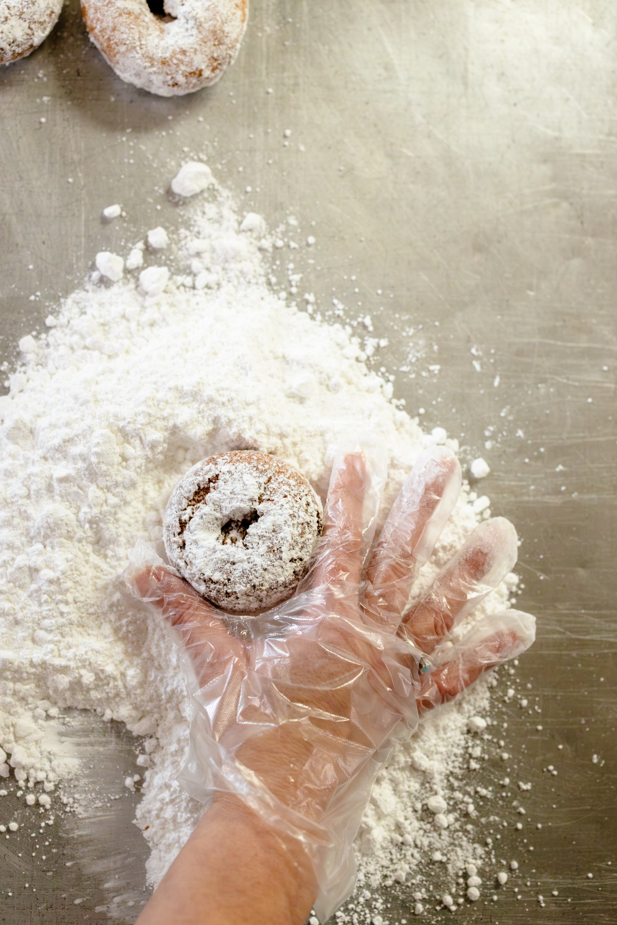 A person with a plastic glove holding a powdered sugar-dusted donut over a flour-covered surface with more donuts in the background.