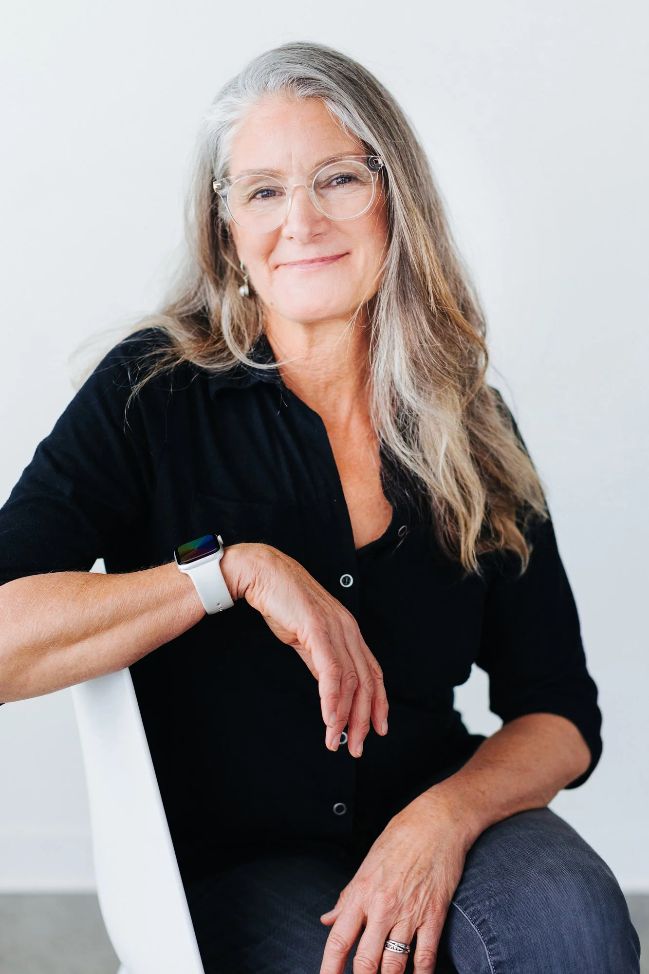 A woman with long gray hair wearing glasses and a black shirt, sitting on a white chair against a plain white background.