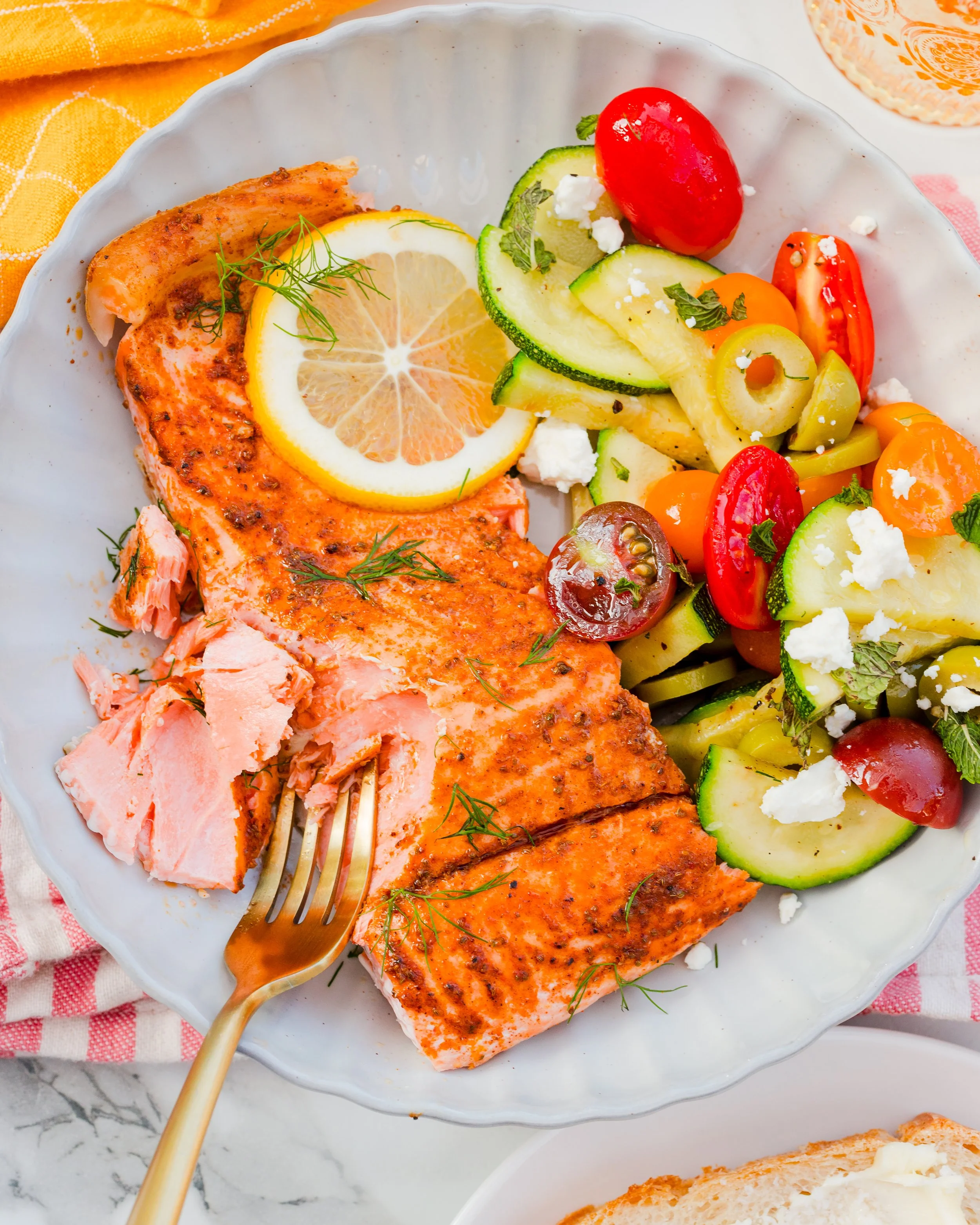 Grilled salmon fillet with lemon slices, served with a fresh vegetable salad containing cucumbers, cherry tomatoes, olives, and feta cheese.