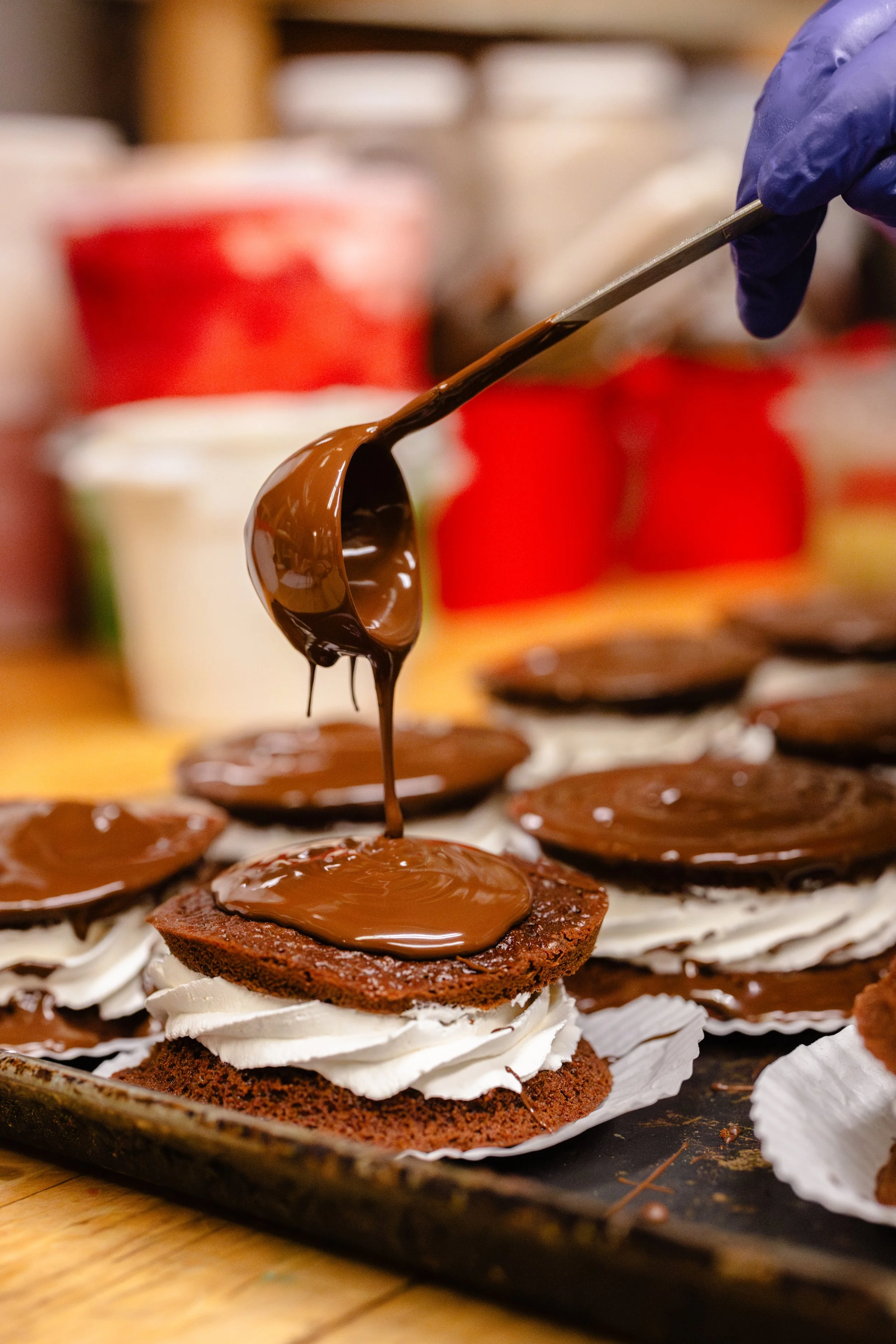 A person wearing a purple glove drizzling melted chocolate onto a layered dessert with whipped cream between two cake layers, on a baking sheet.