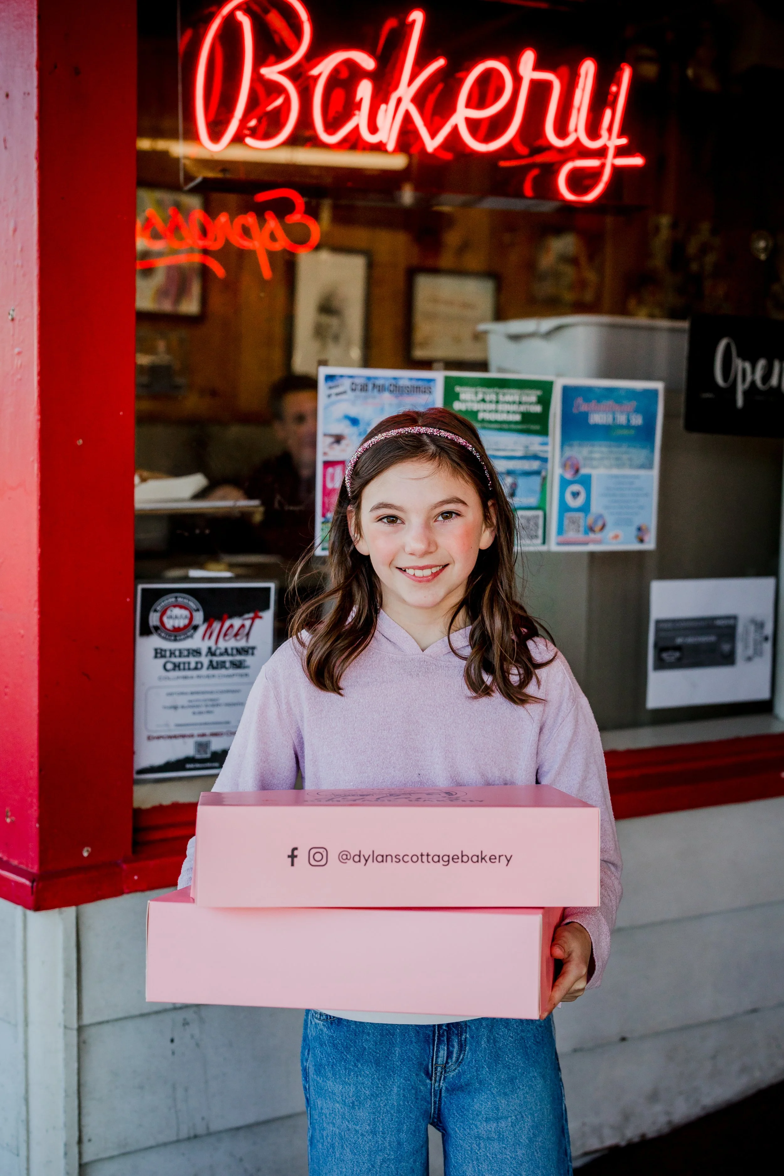 Girl smiling and holding two pink bakery boxes outside a bakery with a neon 'Bakery' sign in the window.