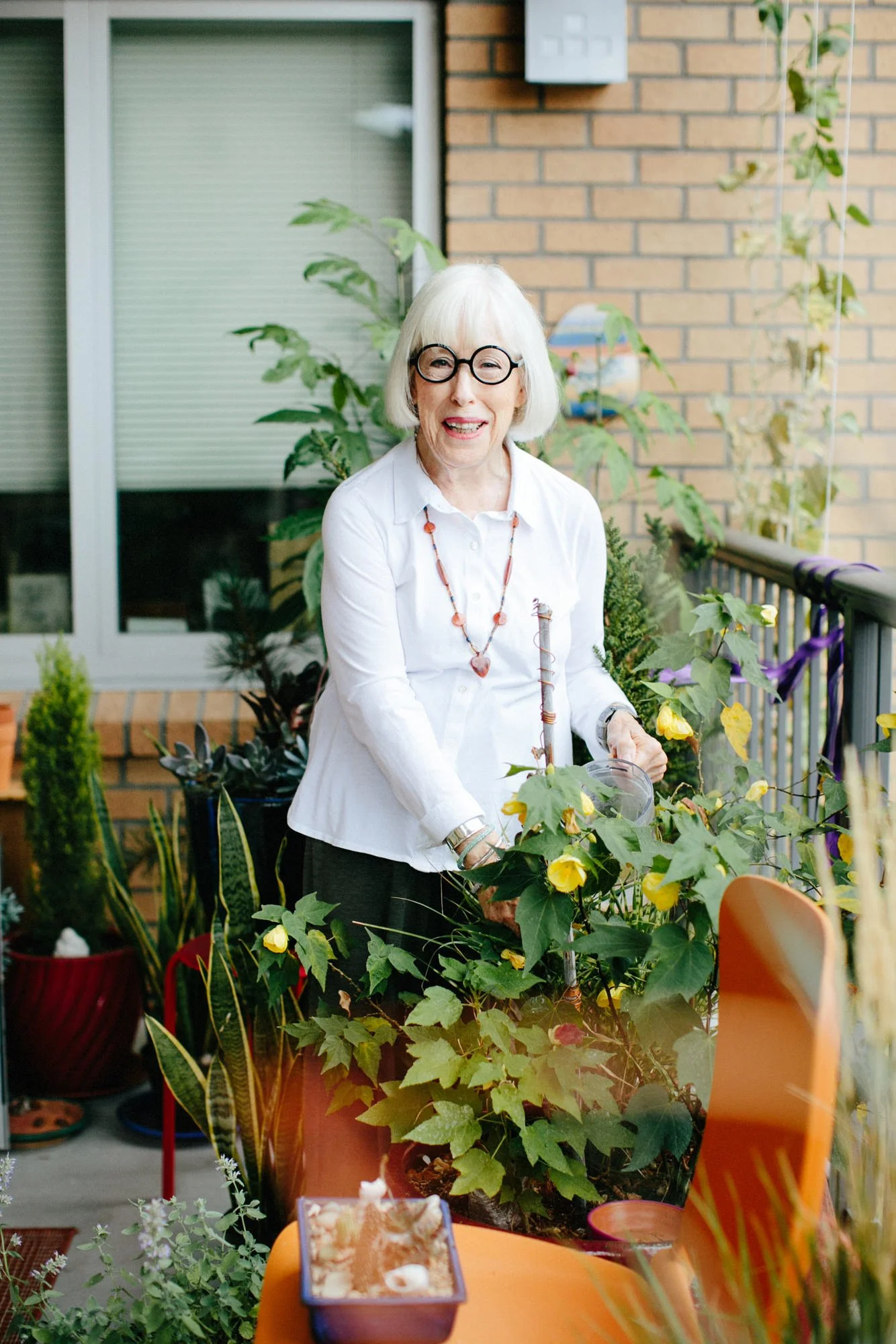 An elderly woman with white hair, glasses, and a white blouse watering plants on a balcony filled with green foliage and colorful pots.