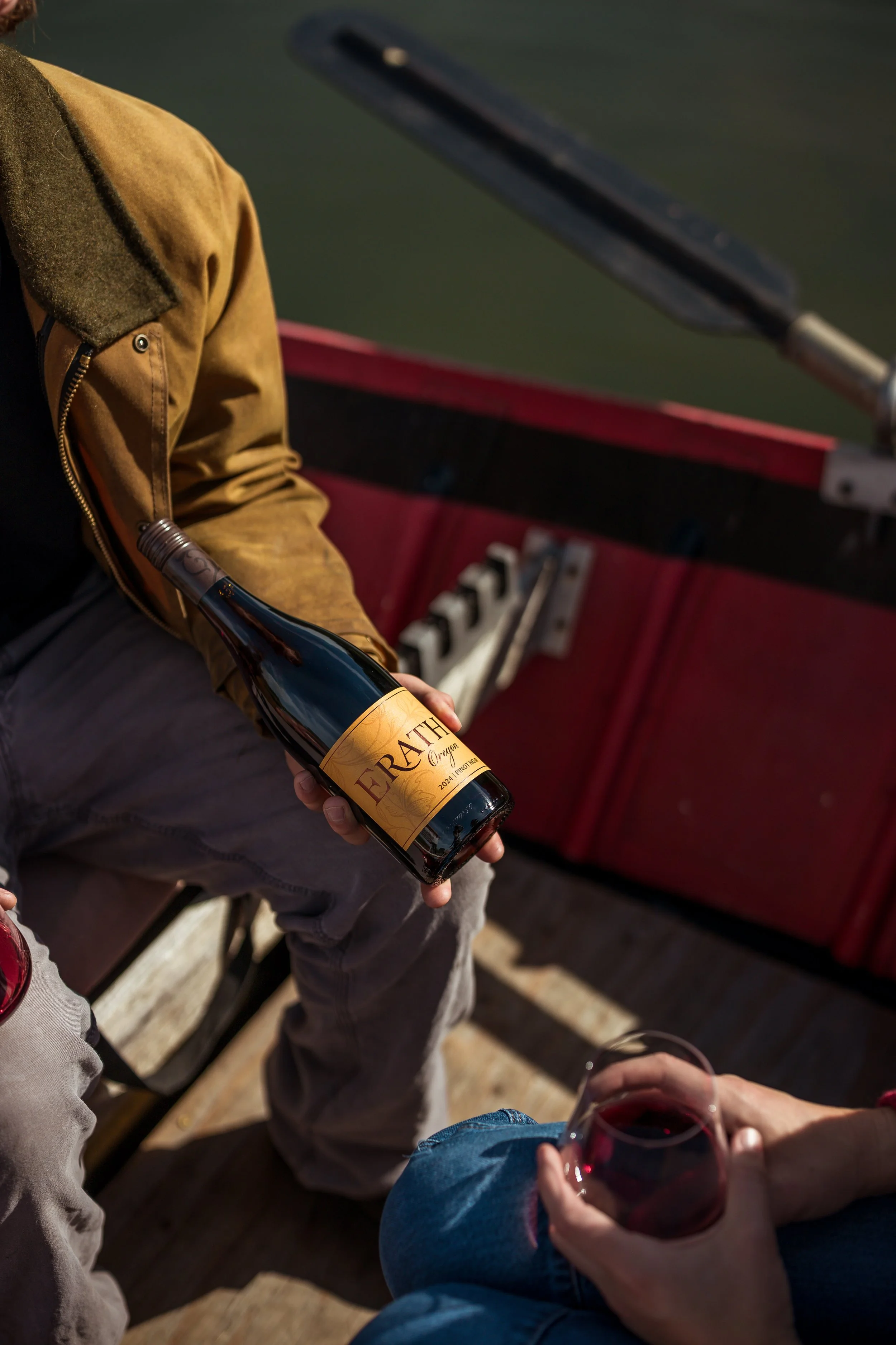A person sits on a boat holding a bottle of Earthy wine, while another person holds a glass of red wine. The boat has a wooden deck and a red railing.