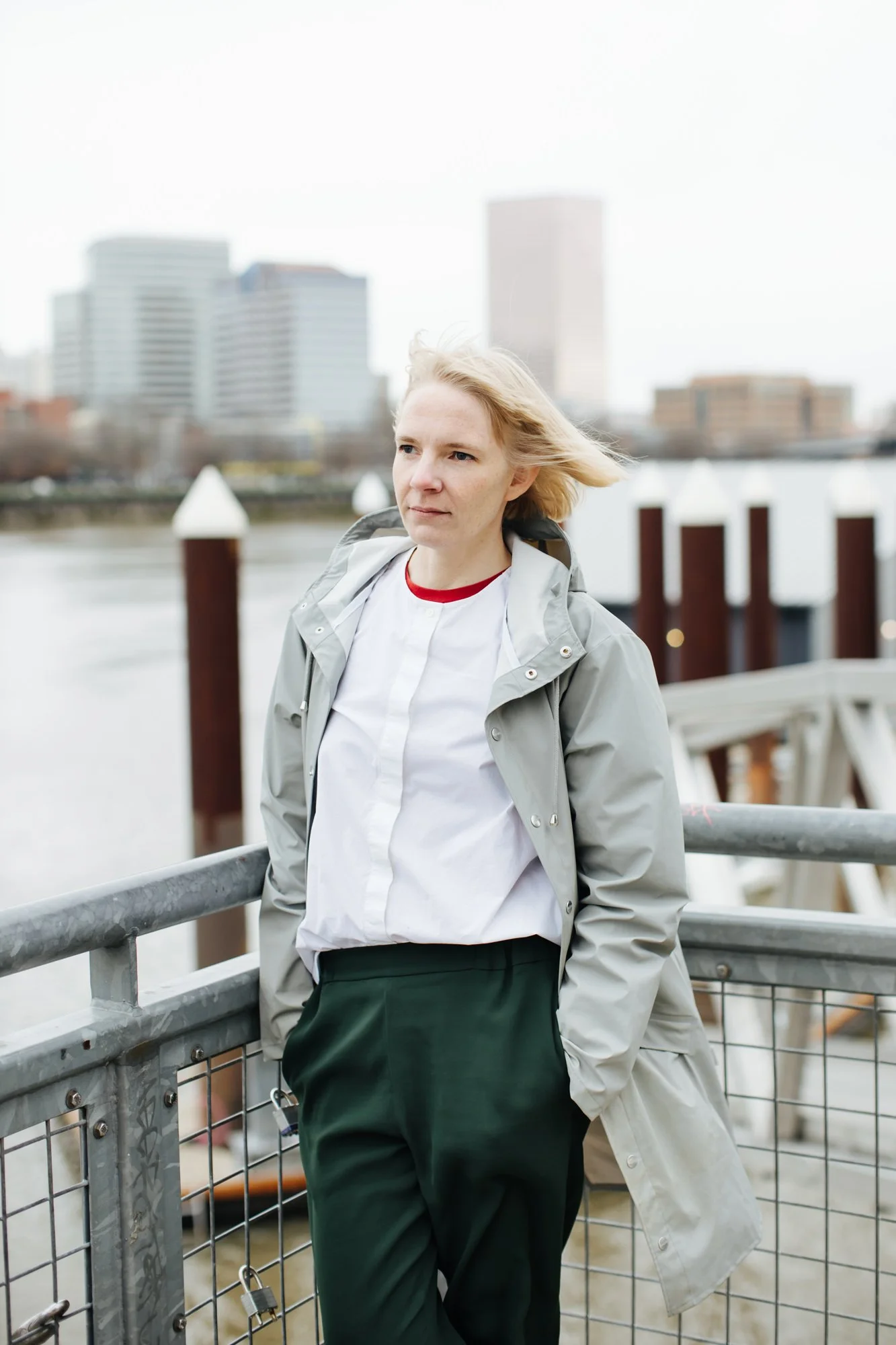 A woman with blonde hair wearing a gray jacket and dark green pants, standing by a railing near a body of water with city buildings in the background.