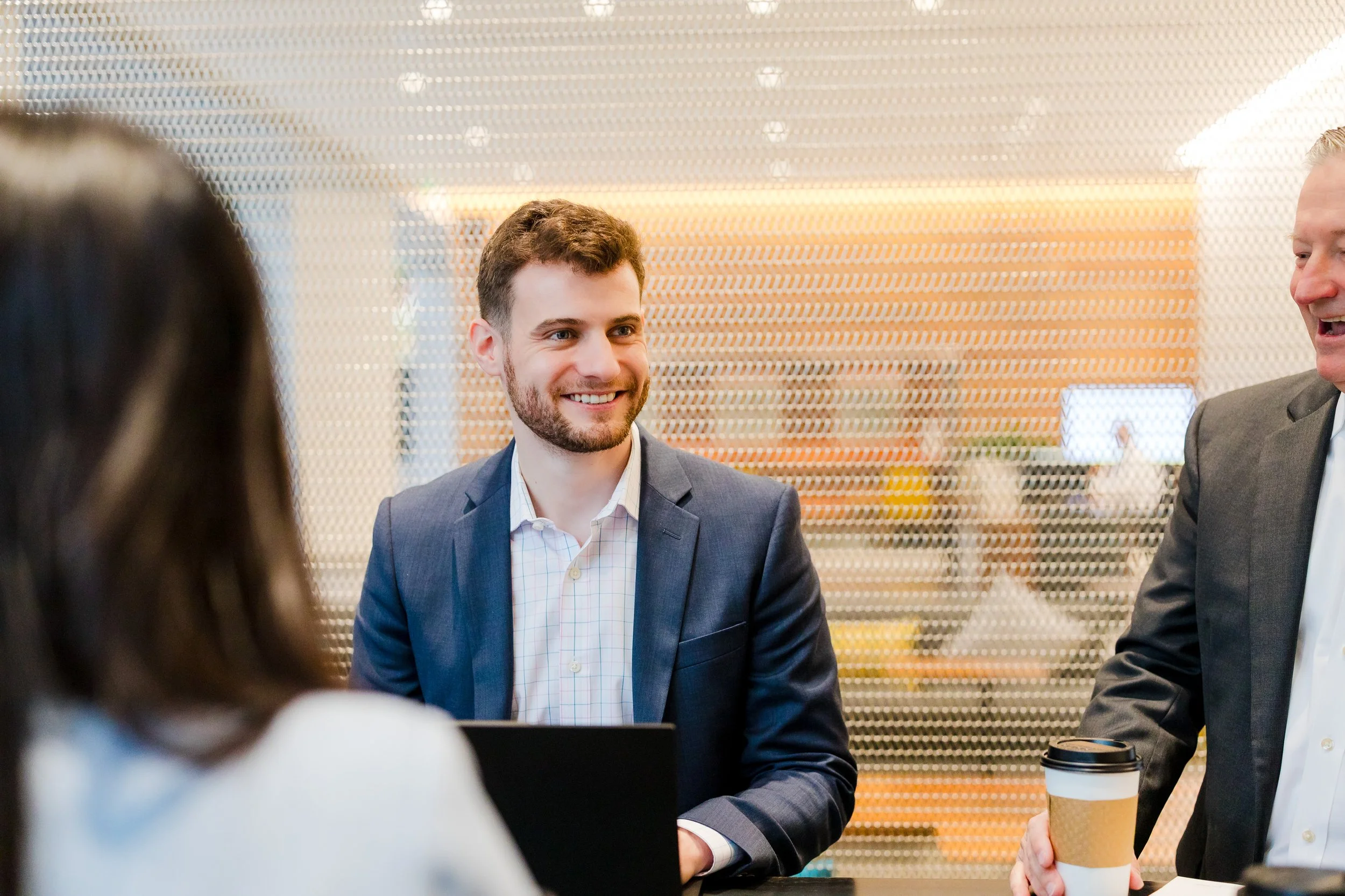 Four business professionals engaging in a conversation in an office setting, with two men in suits smiling and a woman partially visible in the foreground.