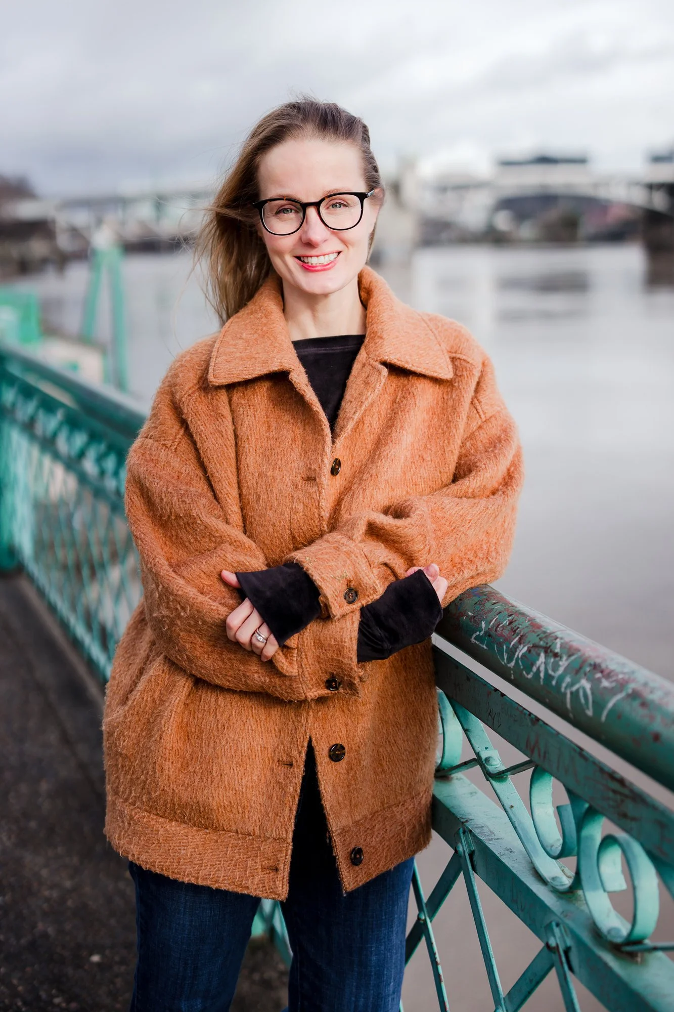 A woman with glasses and long hair, smiling, wearing an orange coat, standing on a bridge over a body of water with boats and a cityscape in the background.