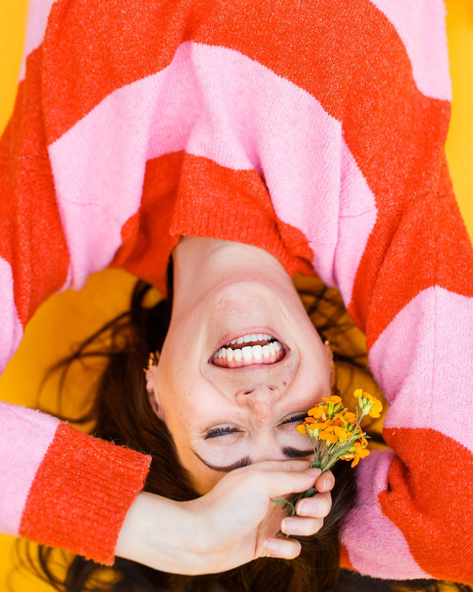 A woman lying on a yellow background, smiling, holding a small bouquet of yellow flowers near her forehead, wearing a red and pink striped sweater.