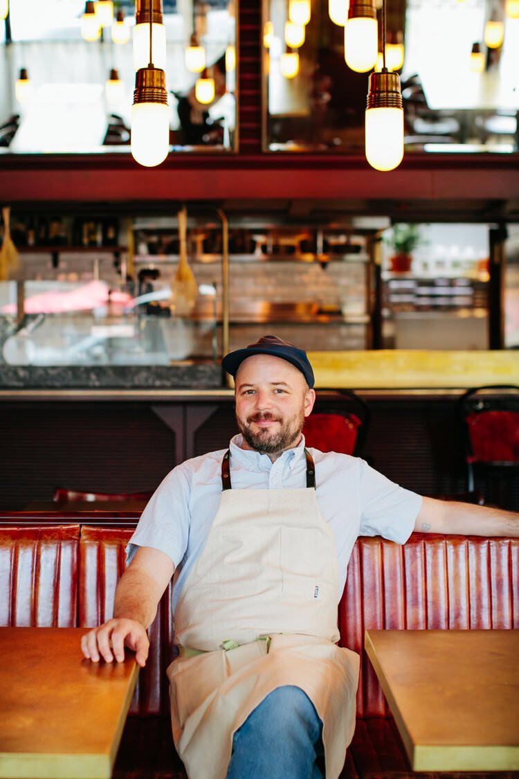A male restaurant worker with a beard, wearing a black cap, white shirt, and beige apron, sitting on a red leather booth inside a restaurant with modern lighting and warm decor.
