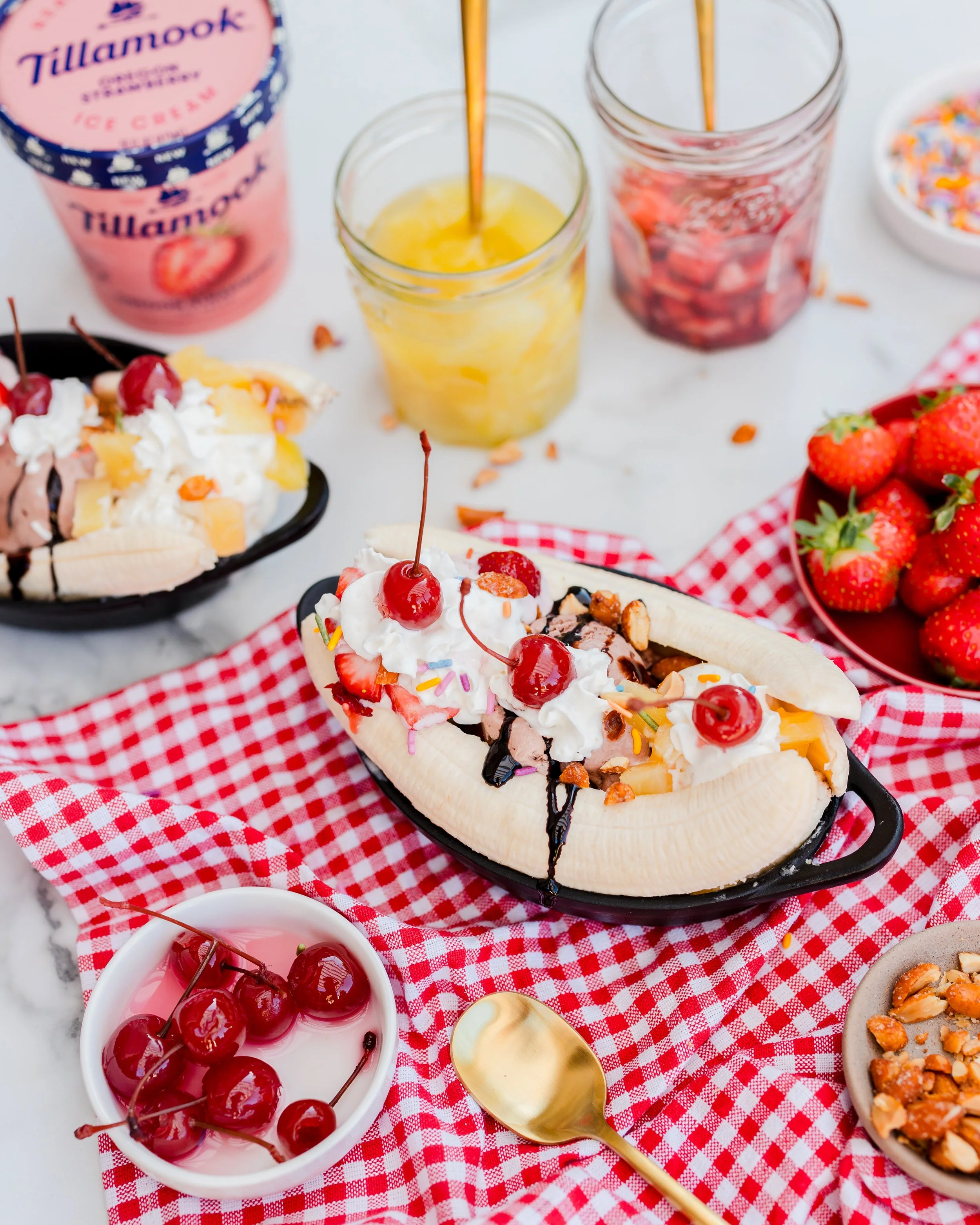 Banana split with whipped cream, cherries, sprinkles, and chocolate syrup, surrounded by strawberries, popsicles, and bowls of cherries and nuts on a red checkered tablecloth.