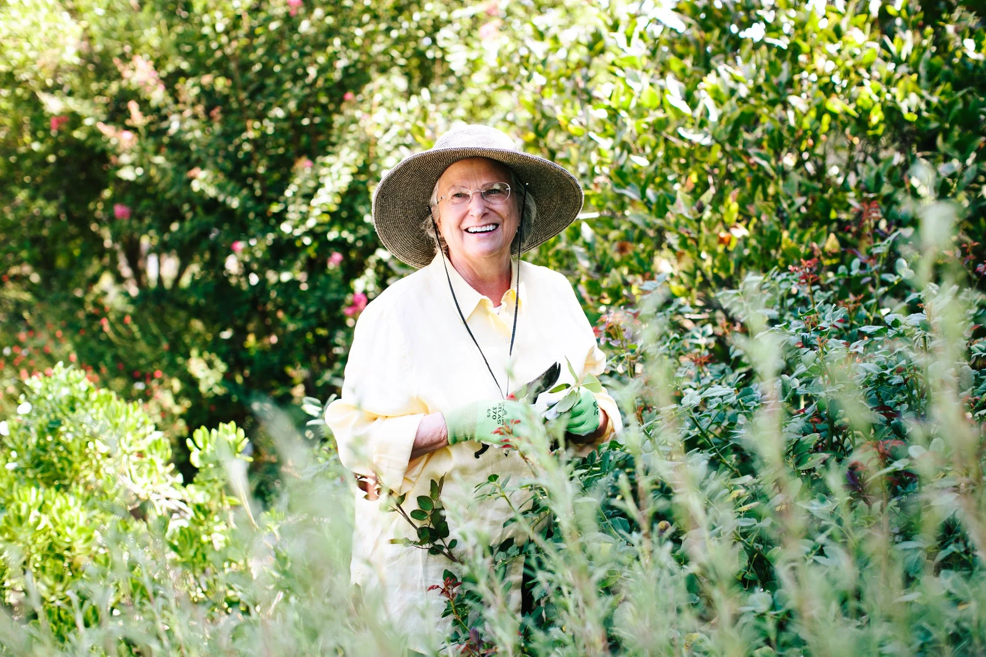 An elderly woman with glasses, a wide-brimmed hat, and green gardening gloves, smiling while standing among bushes on a sunny day.