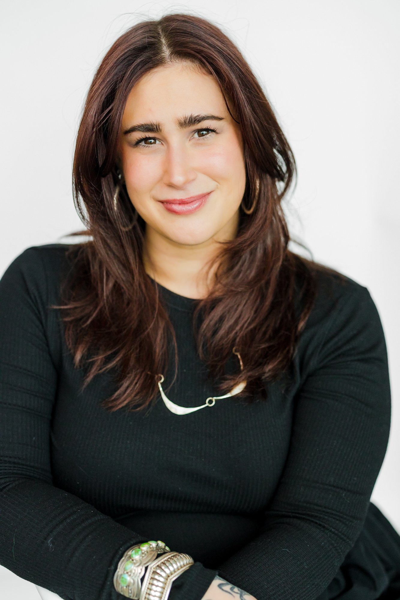 A woman with shoulder-length dark red hair, wearing a black long-sleeve top, gold hoop earrings, a silver necklace, and bracelets, smiling at the camera against a white background.
