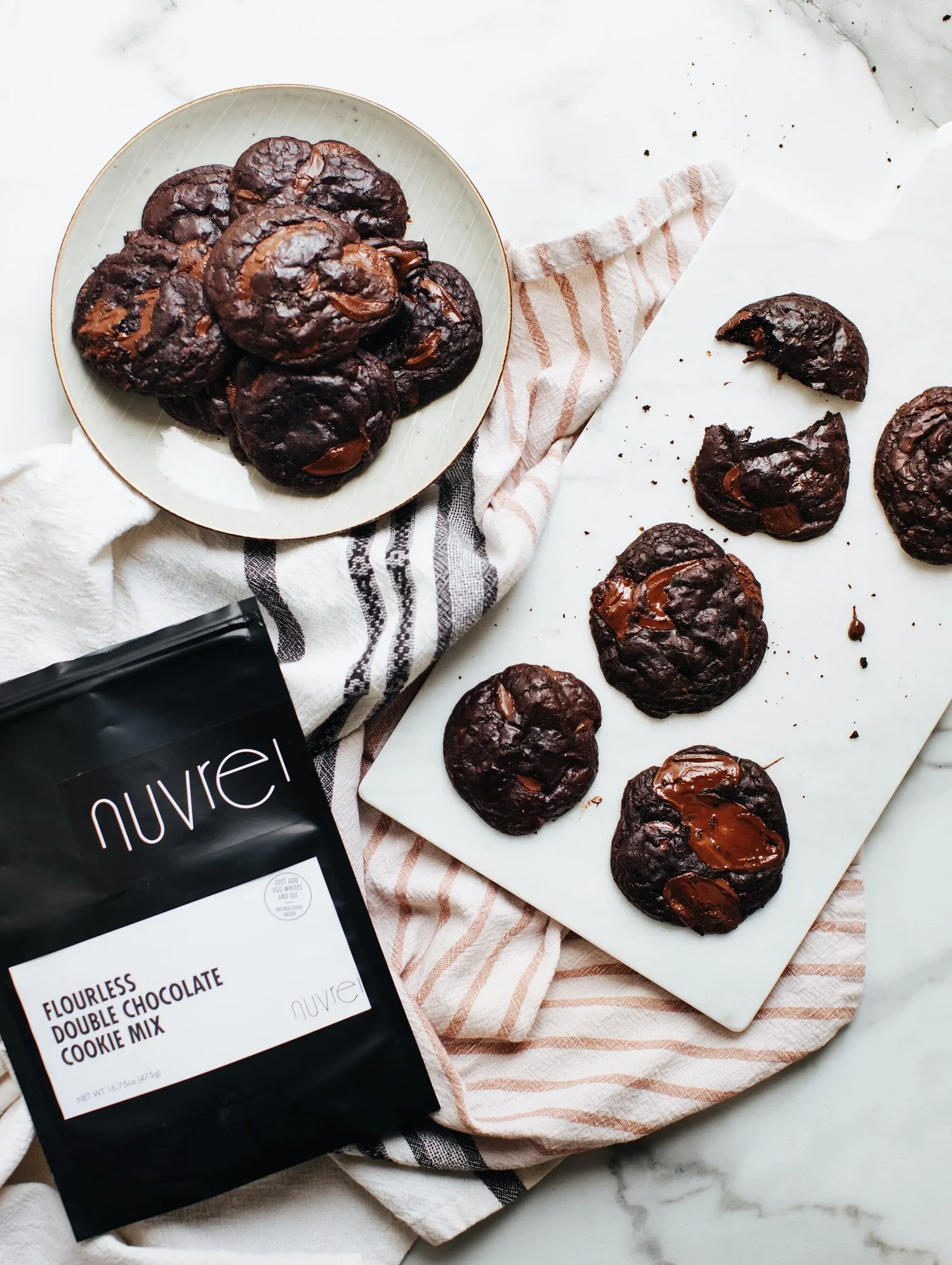 Chocolate cookies on a white platter and a marble surface, next to a package of Nuve flourless double chocolate cookie mix. Some cookies are broken on the marble surface.