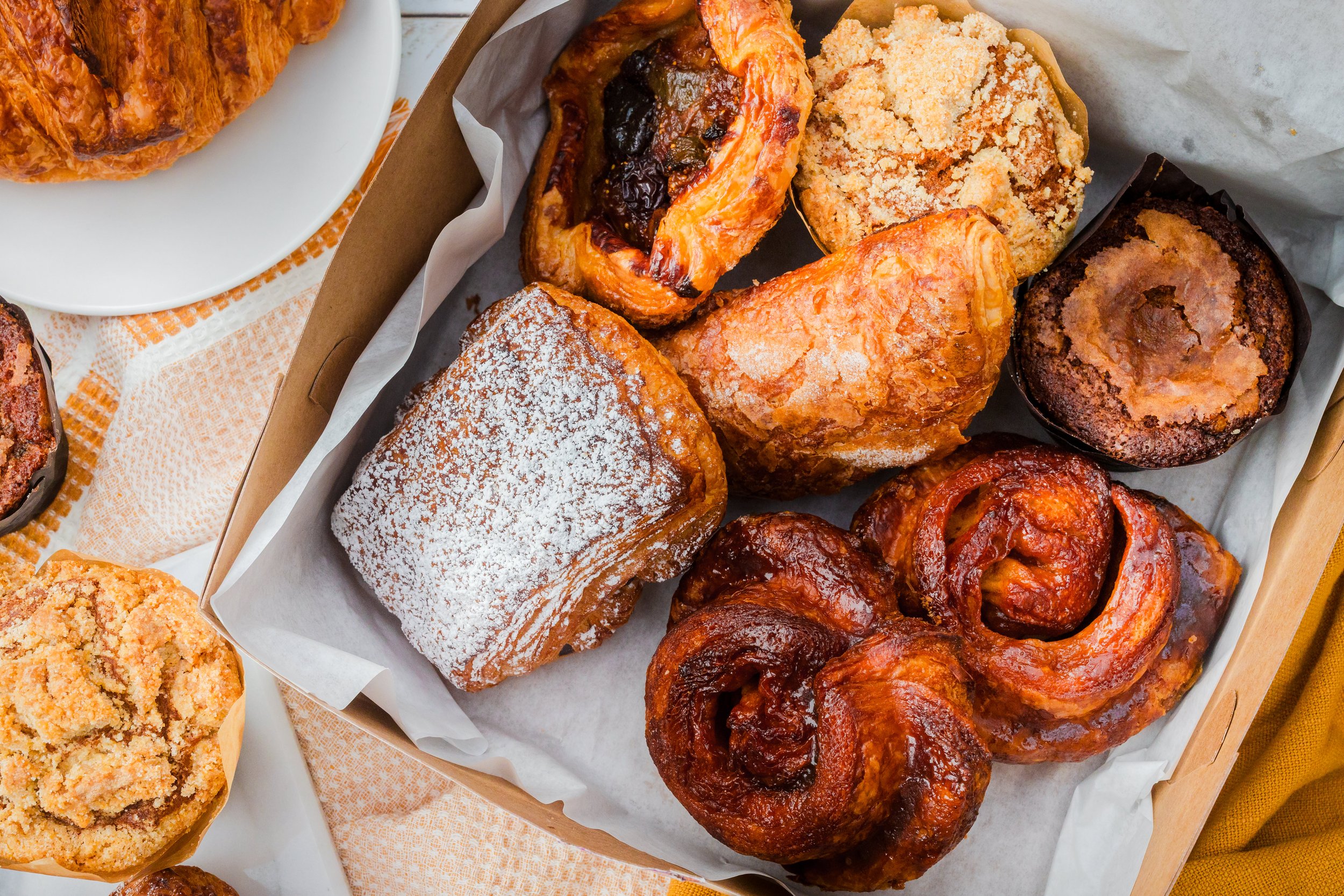 Assorted baked goods including cinnamon rolls, muffins, and pastries in a cardboard tray lined with paper