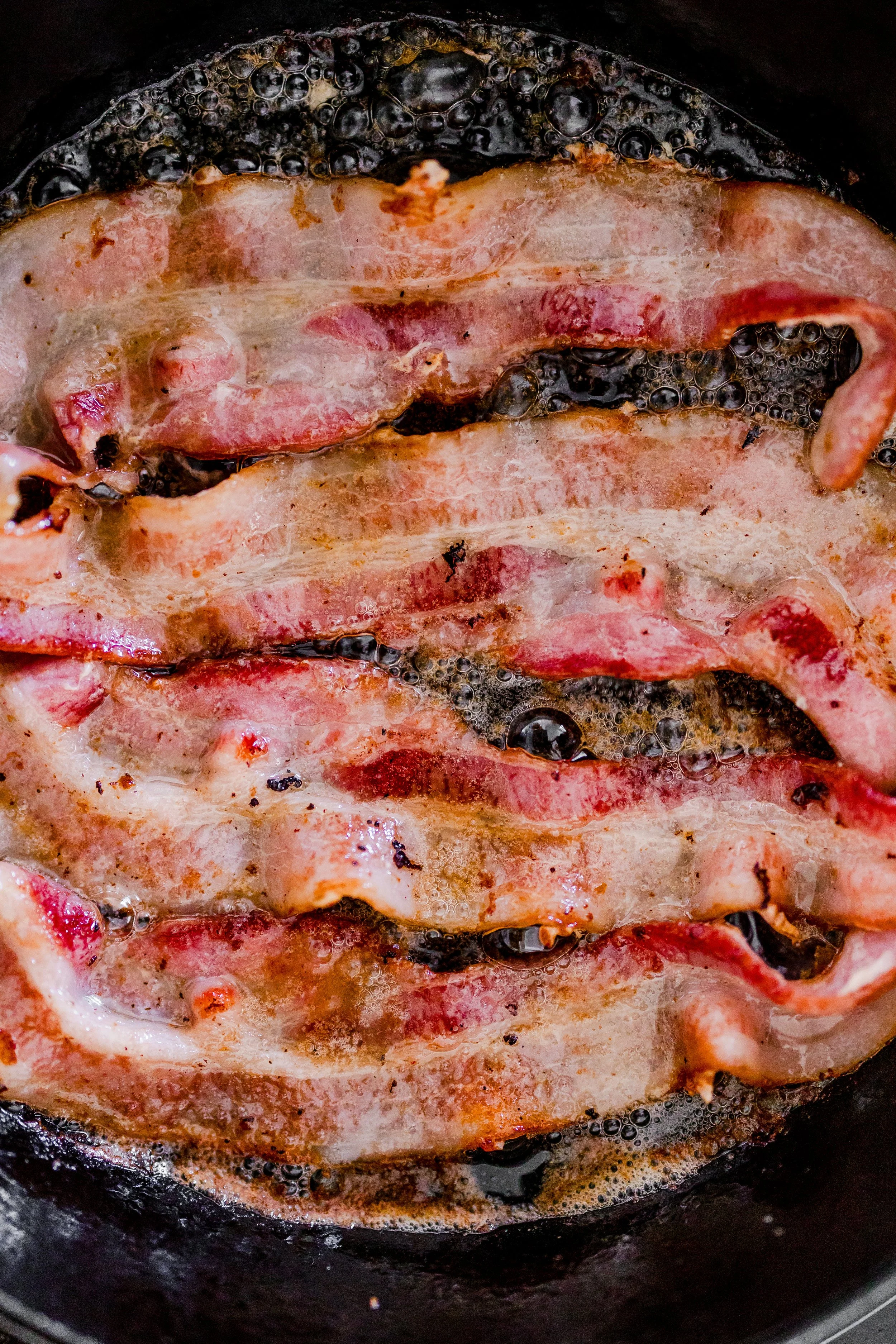 Close-up of bacon frying in a skillet, with bubbling oil.