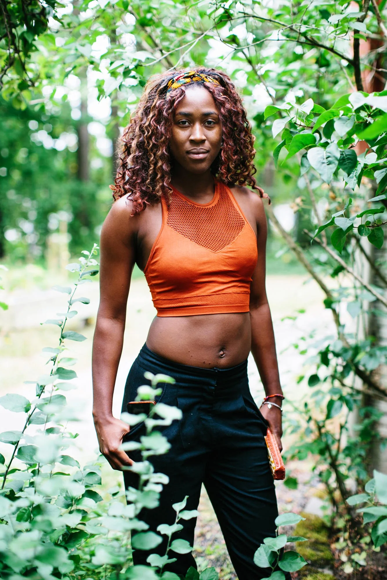 A woman with curly reddish-brown hair standing outdoors surrounded by green foliage, wearing an orange crop top and black pants.