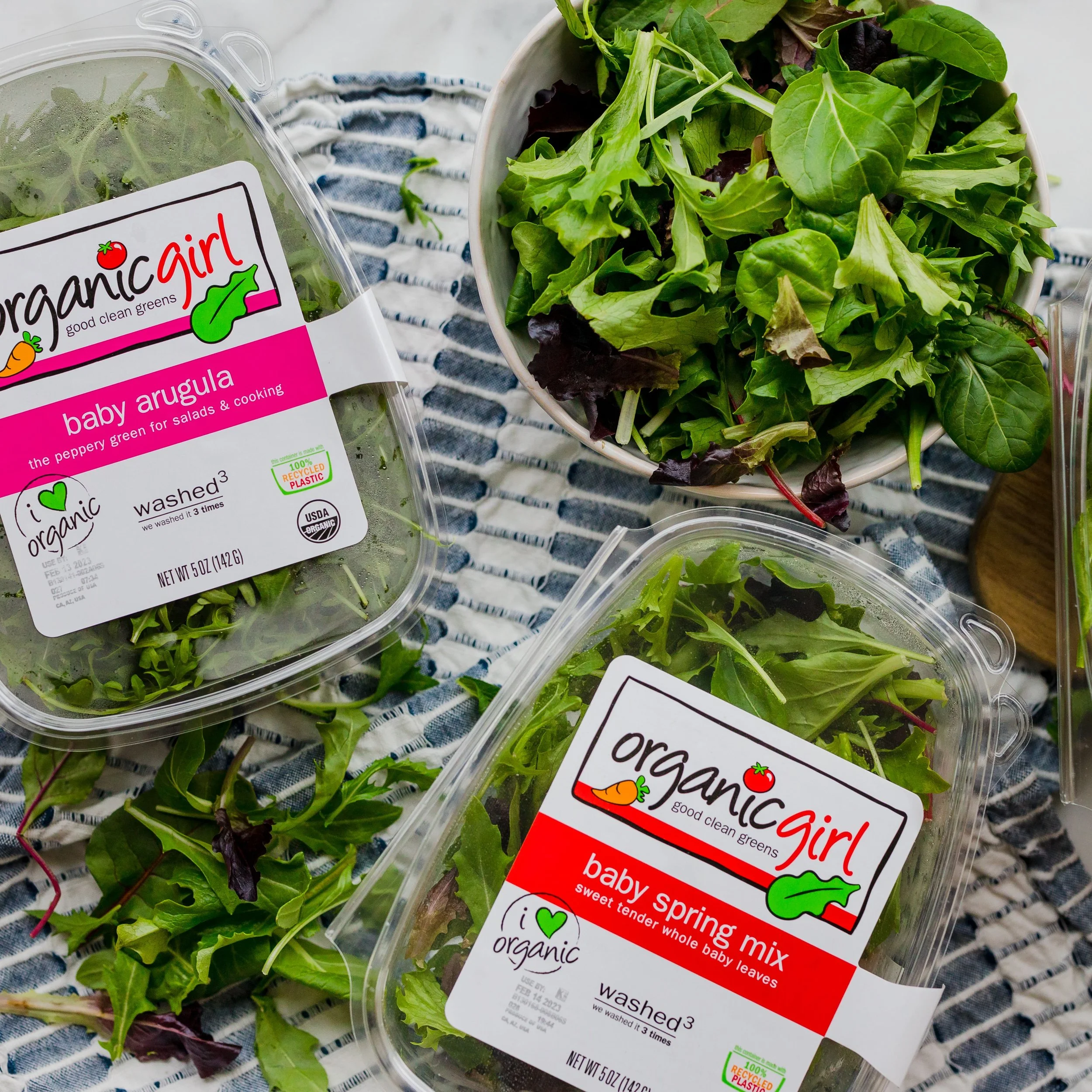 Containers of organic baby arugula and organic baby spring mix with a bowl of loose mixed greens on a striped cloth surface.