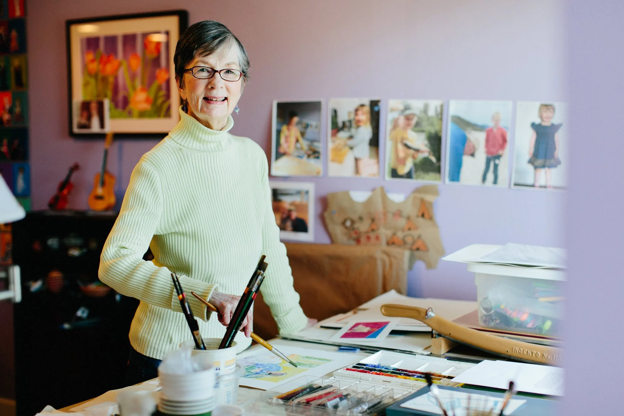 An elderly woman with short gray hair and glasses, wearing a yellow turtleneck sweater, standing at a table with art supplies in a colorful room. Behind her, there are framed pictures of children on the purple wall and a brown couch with a decorative