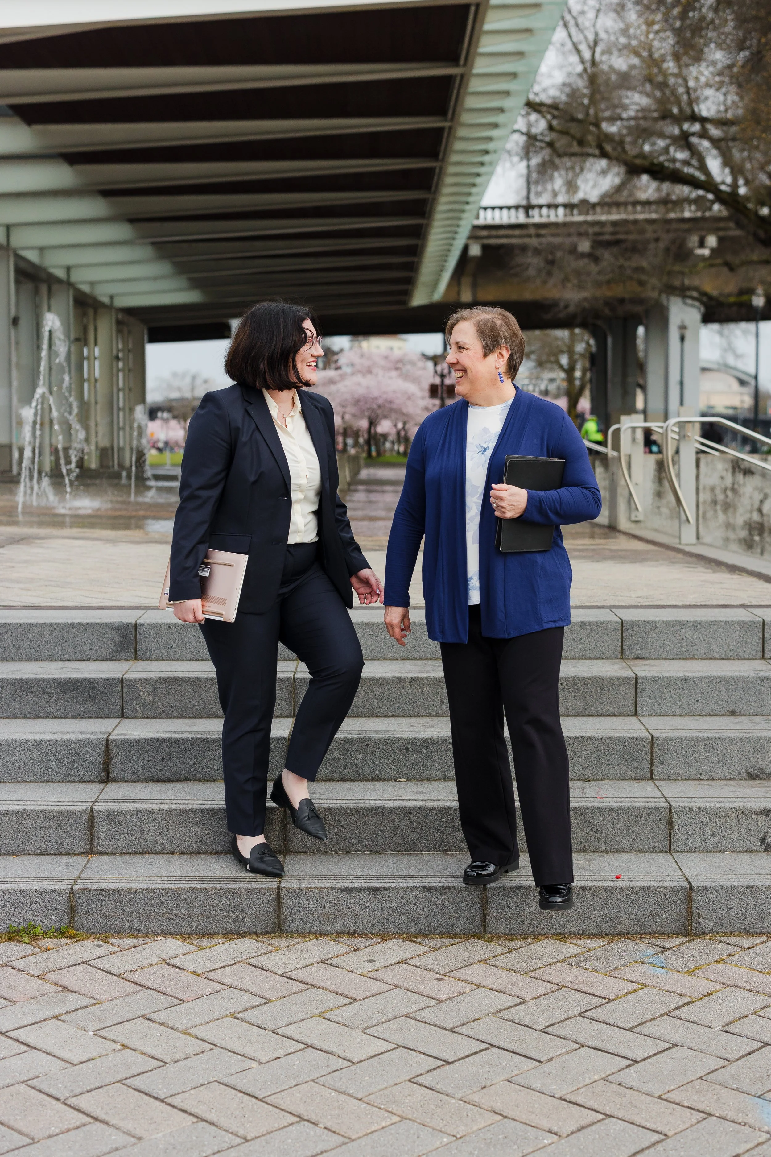 Two women in professional attire talking and smiling on outdoor steps, holding folders, with trees and a bridge in the background.