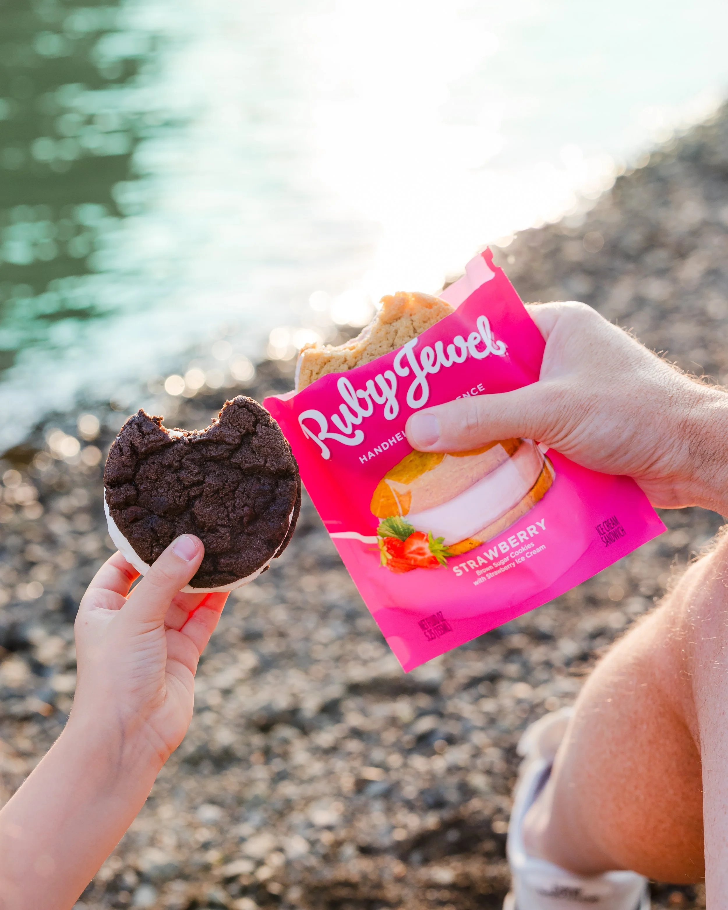 A person holding a RubyJewel strawberry ice cream sandwich in front of a rocky shoreline and water, with another person holding a cookie in their hand. The ice cream sandwich packaging is pink.