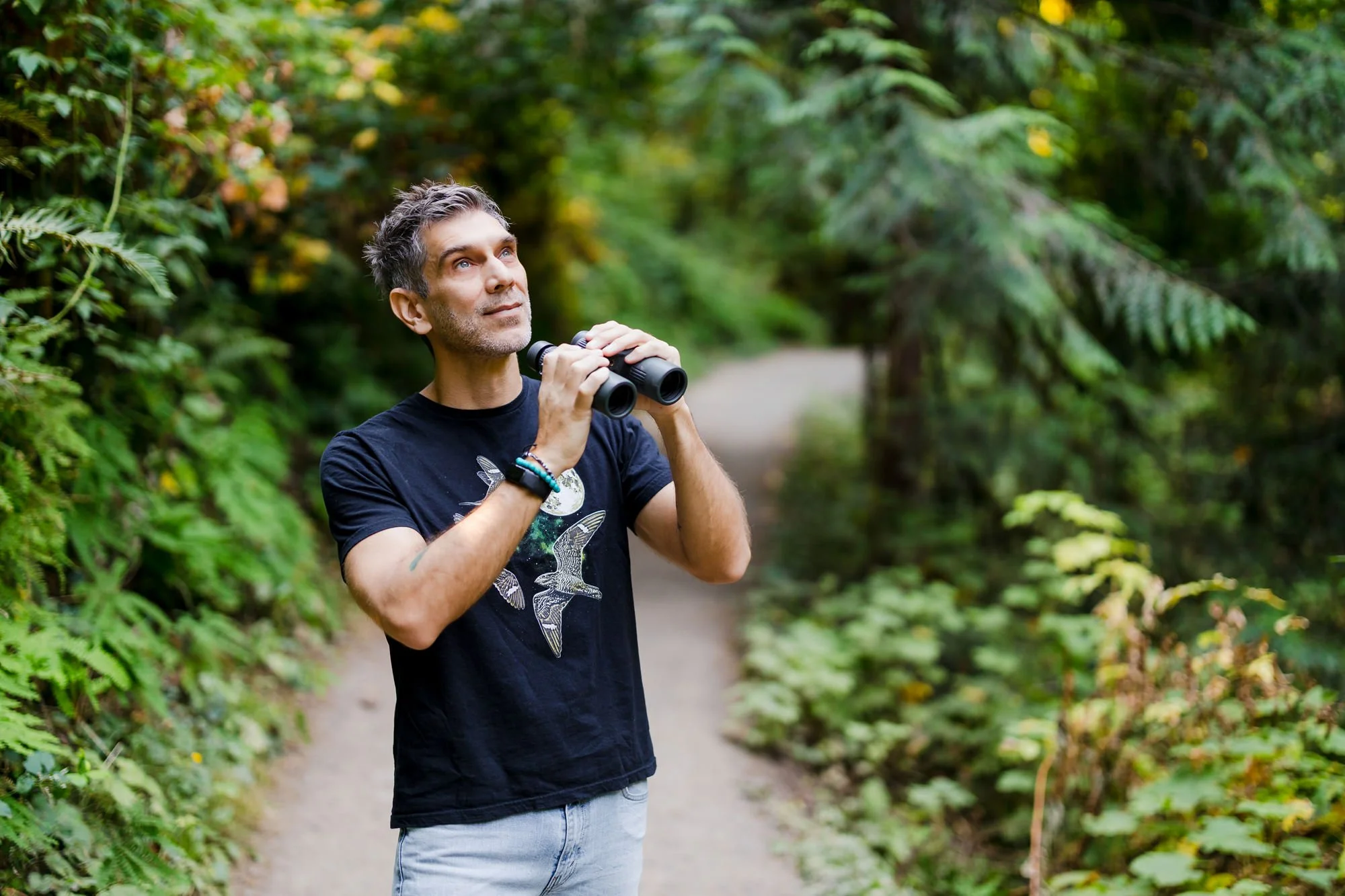 A man with gray hair and beard looking up while holding binoculars in a forested area.