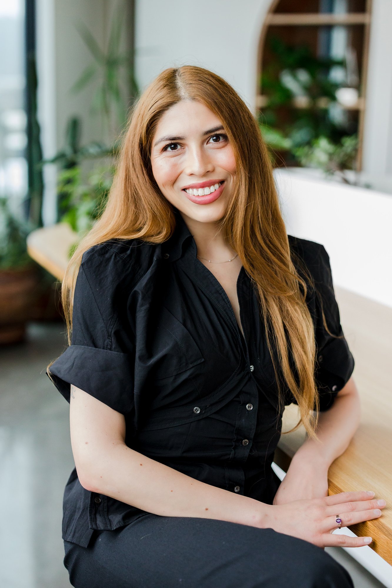 A young woman with long, red hair sitting at a wooden counter in a bright, modern space. She is smiling and wearing a black shirt, with some plants and shelving in the background.