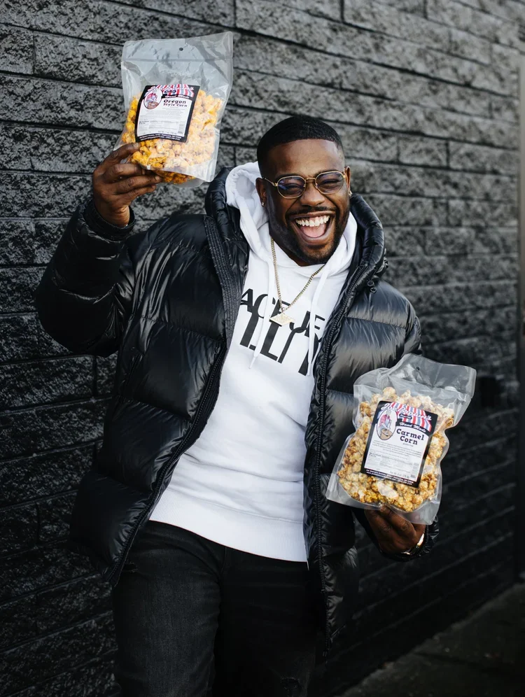 A smiling man wearing glasses, a black puffer jacket, and a white hoodie holding two bags of caramel popcorn, standing against a black brick wall.