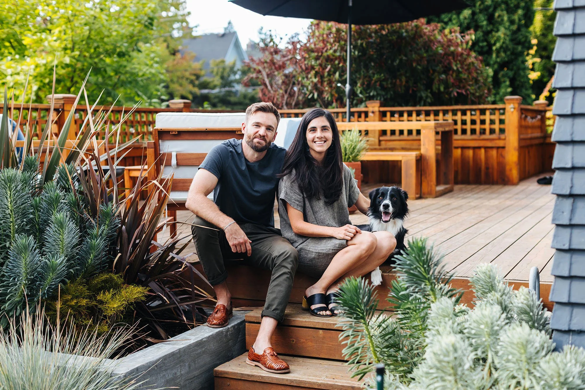 A man, woman, and dog sitting on a wooden deck outdoors with plants and trees in the background.