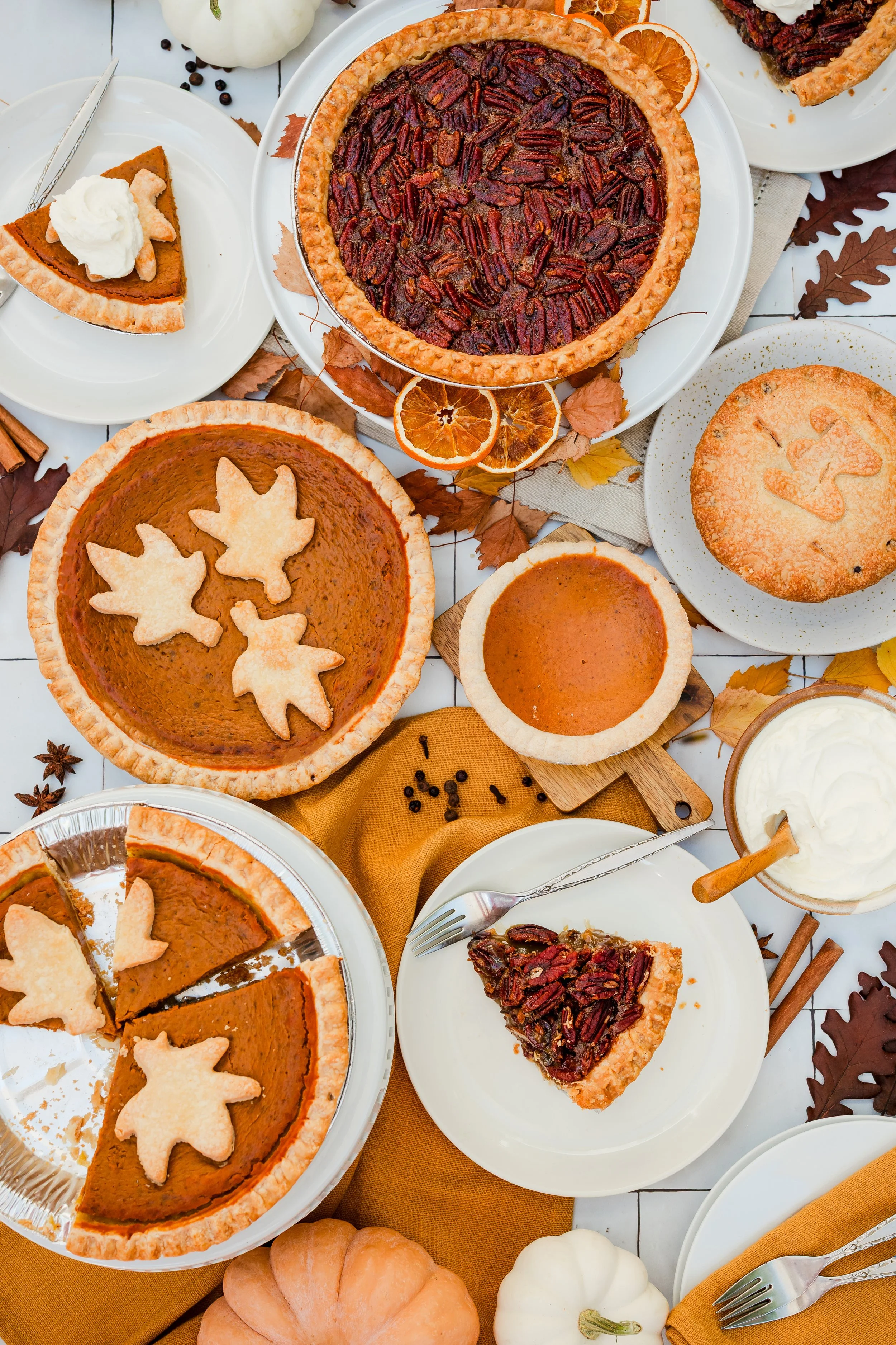 An assortment of fall-themed pumpkin and pecan pies on a white table, decorated with orange and brown leaves, dried oranges, and pumpkins, accompanied by whipped cream and slices.