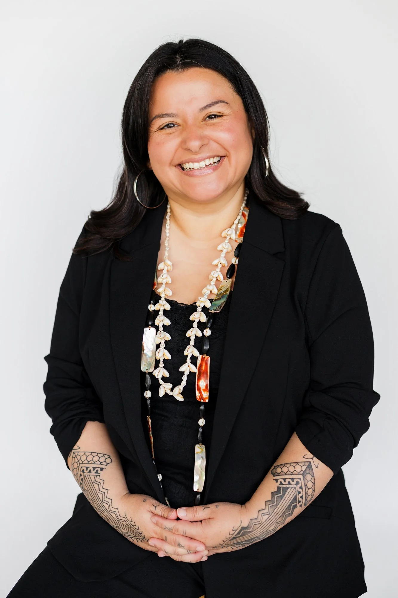 A woman with dark hair, wearing a black blazer, large hoop earrings, and layered necklaces, sitting against a plain white background and smiling at the camera.
