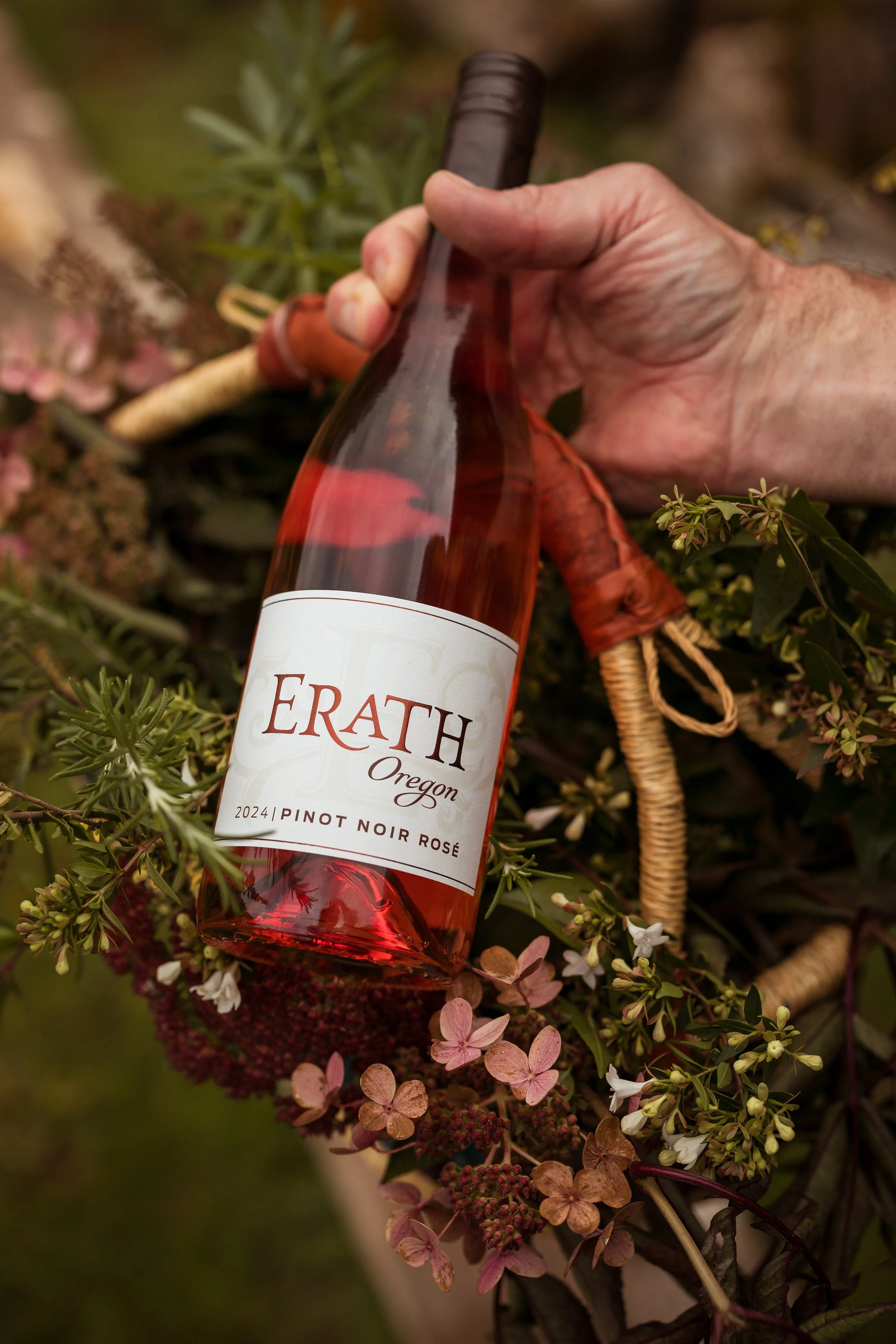 A hand holding a bottle of Erath Oregon Pinot Noir Rosé wine with a label indicating 2024 vintage, surrounded by flowers and leaves.