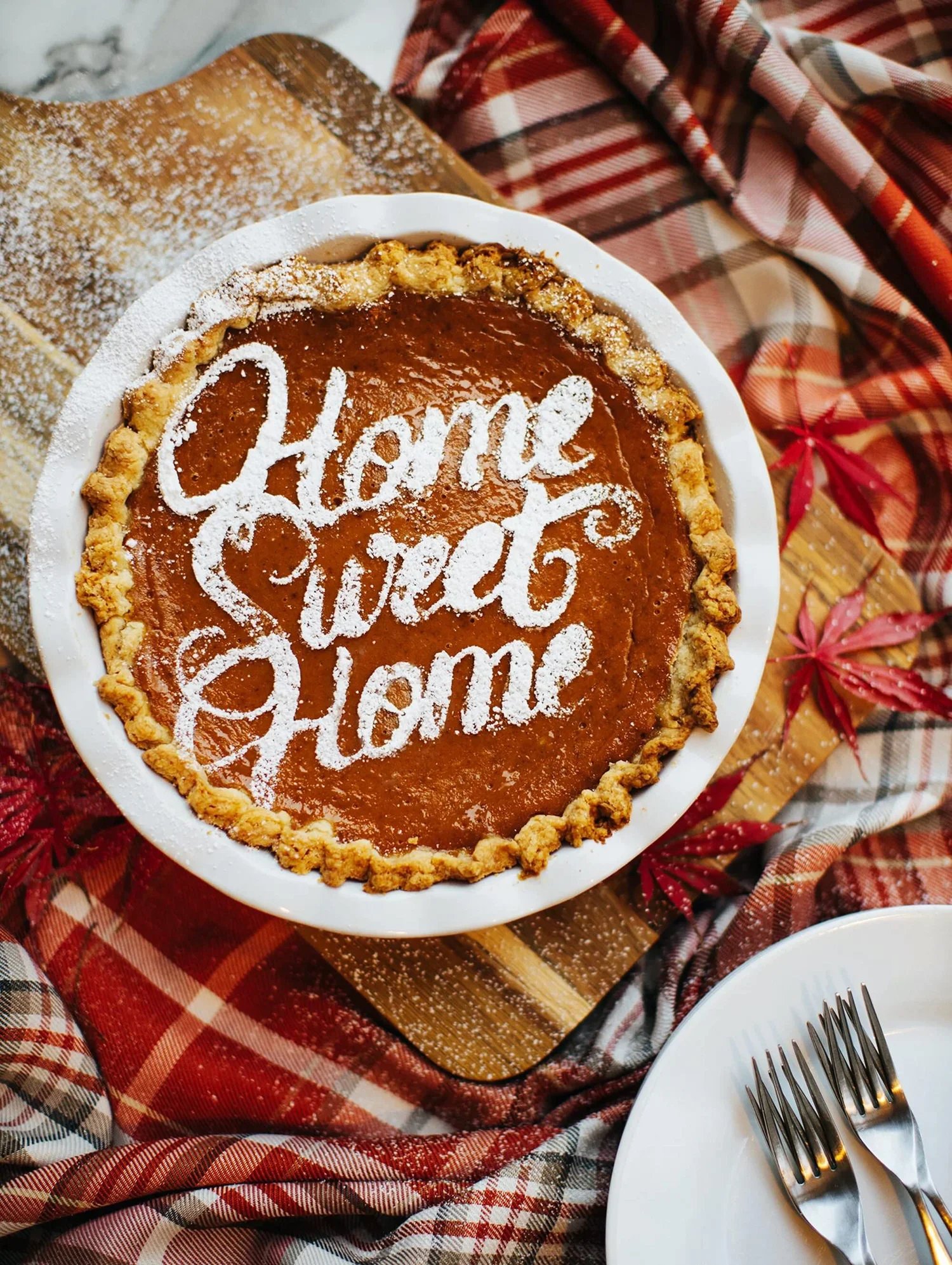 Pumpkin pie with 'Home Sweet Home' written in powdered sugar on top, surrounded by autumn leaves and a plaid cloth.