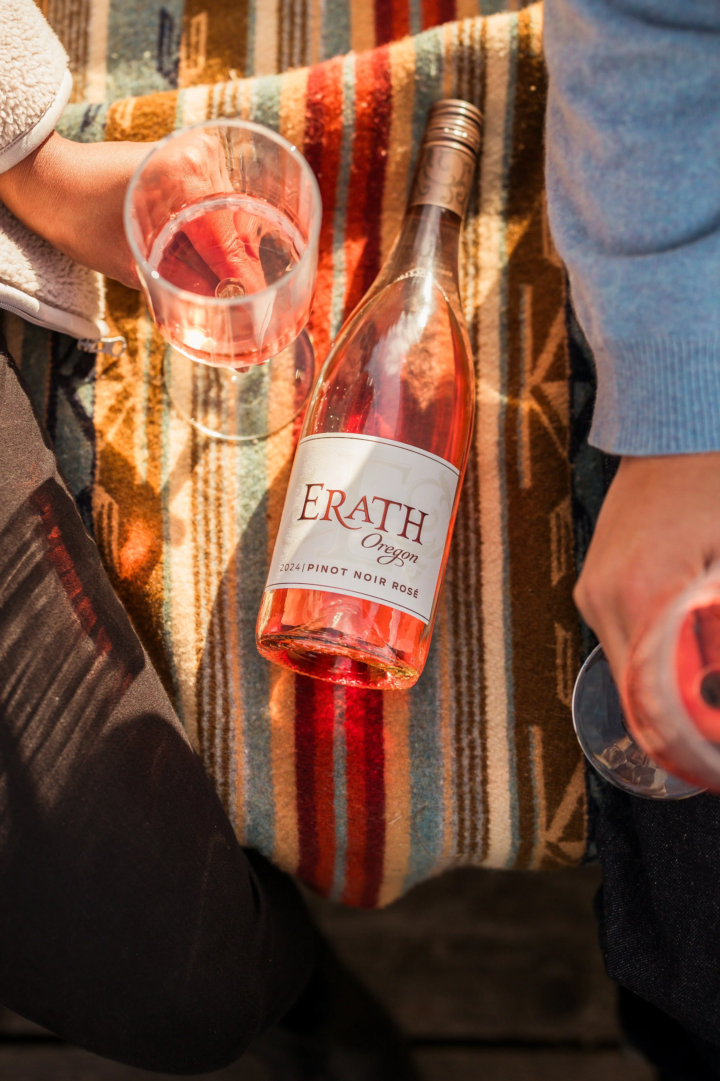Two people holding glasses of Rosé wine, sitting on a colorful striped blanket with a bottle of Erath Oregon Pinot Noir Rosé between them.