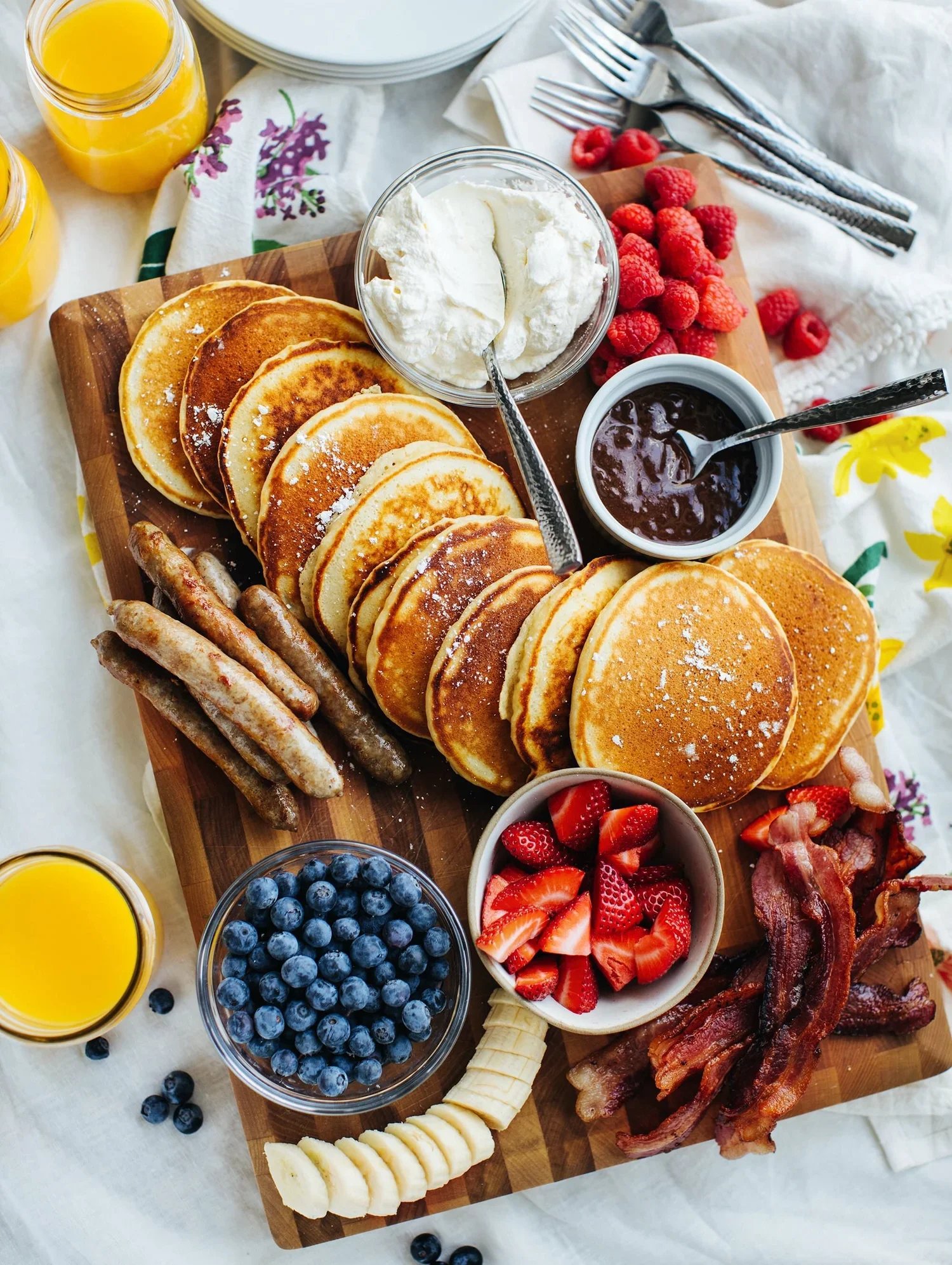 A wooden tray with pancakes, bacon, sausage links, strawberries, blueberries, banana slices, whipped cream, chocolate sauce, raspberry jam, orange juice, and raspberries.