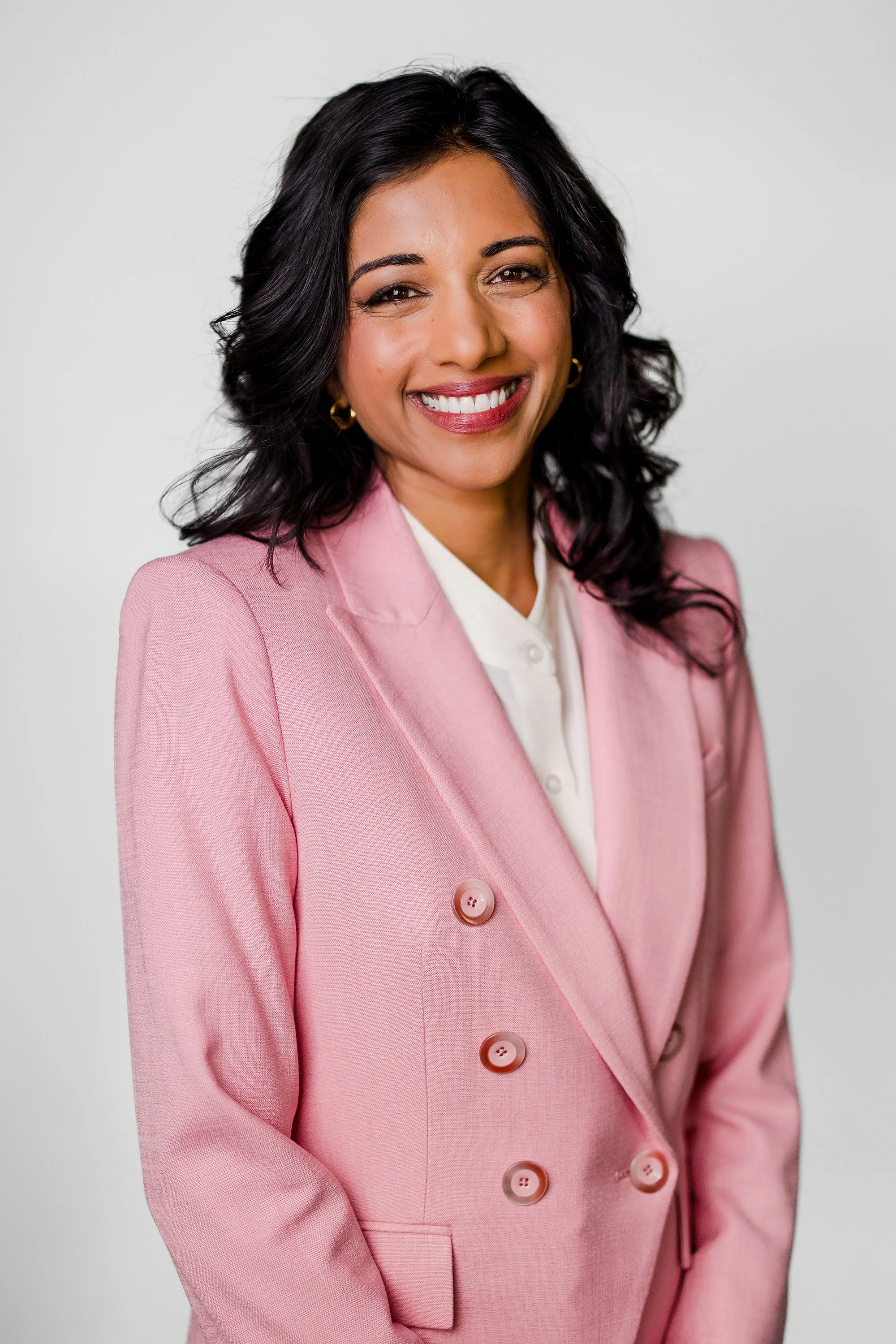 A woman with black wavy hair, wearing a pink blazer with buttons, a white blouse, and gold hoop earrings, smiling against a plain white background.
